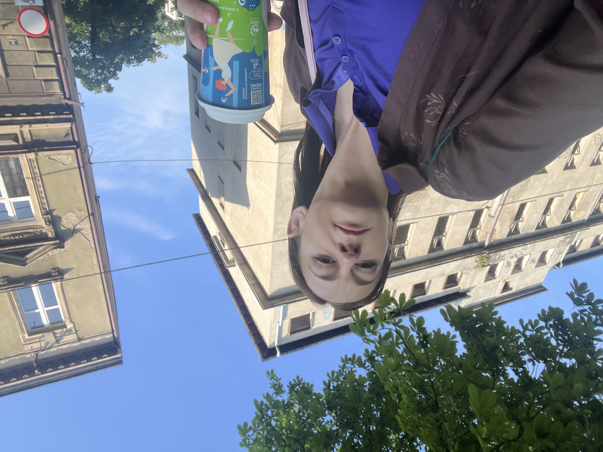 A woman is taking a selfie outdoors in front of two buildings. She is holding a coffee cup in her right hand and is wearing a purple polo shirt and a brown jacket. The buildings appear to be old and are weathered. The sky is clear and blue. The woman's face is in the foreground, and she is looking towards the camera. The coffee cup has a blue and white design with images of people dancing. There are trees and foliage visible in the background.