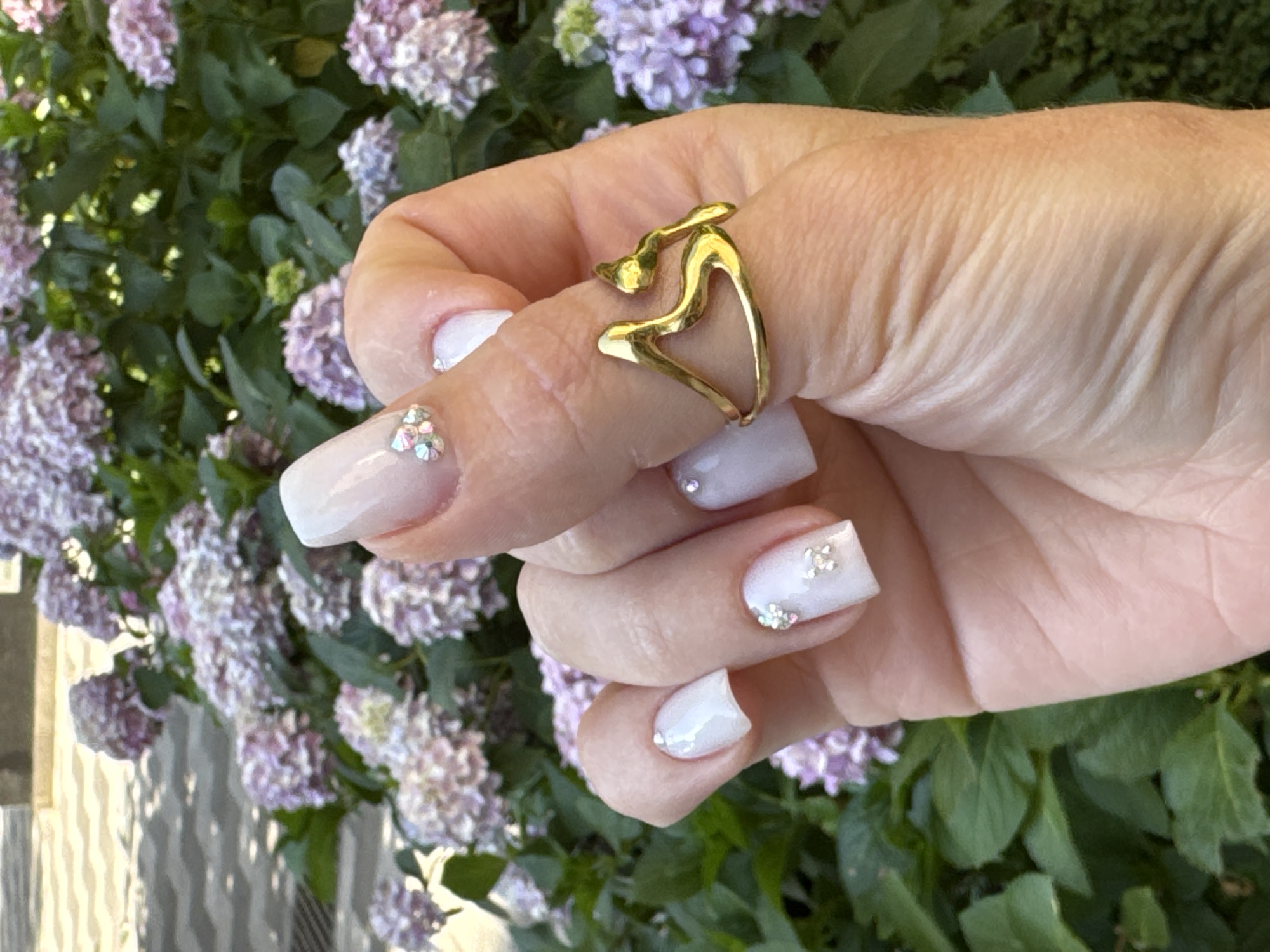 The image shows a close-up of a person's hand adorned with a gold ring and white, square-shaped nails decorated with small rhinestones. The ring is abstract and appears to be made of polished gold. The nails have a French manicure style with a clear or pale pink polish and white tips, some with subtle sparkle. The hand is positioned in front of a blurred background of lush green foliage and clusters of light purple hydrangeas. The lighting suggests it is daytime, possibly outdoors, with soft shadows indicating natural light. The scene evokes a sense of natural beauty and personal adornment. The location context is Budva, Montenegro, but this cannot be visually confirmed from the image alone. There is no visible text in the image.