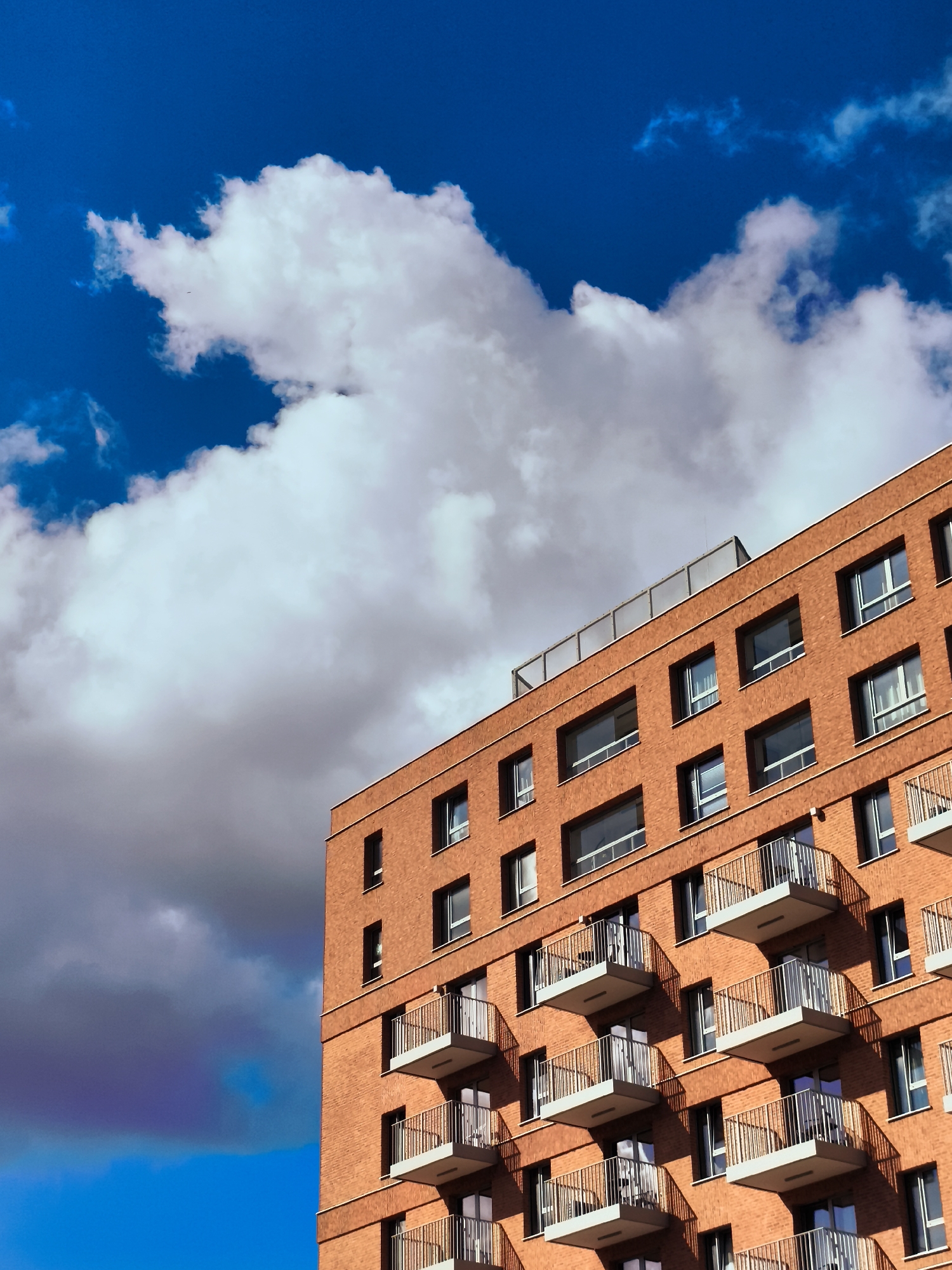 An exterior, medium shot captures a contemporary, multi-story brick building with balconies against a dramatic blue sky filled with white and gray clouds. The building's facade is constructed of reddish-brown bricks with evenly spaced, dark-framed windows. Each level features several balconies with white railing and a cantilevered gray floor. A rooftop edge with a glass barrier is visible at the top of the building. The bright sunlight casts shadows from the balconies onto the brickwork. The image was taken in the city of Gdańsk, Poland.