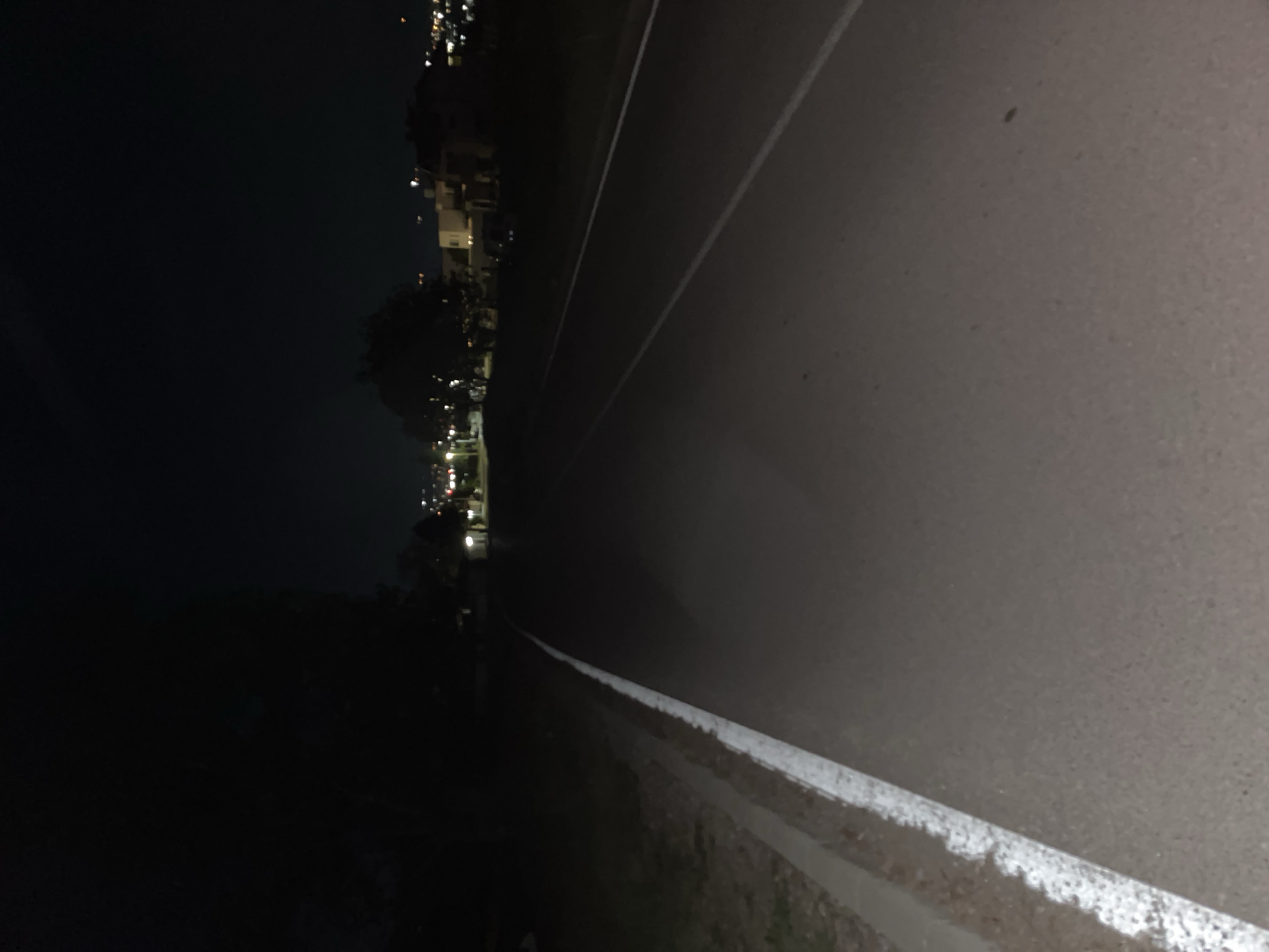 The image depicts an urban scene at night. The right portion shows a grey concrete road surface with white lane markings. To the left, a cityscape with lights visible in the distance and along the horizon is seen. The surrounding area seems dark with little to no illumination, hinting at a sparse or vacant environment next to the road.