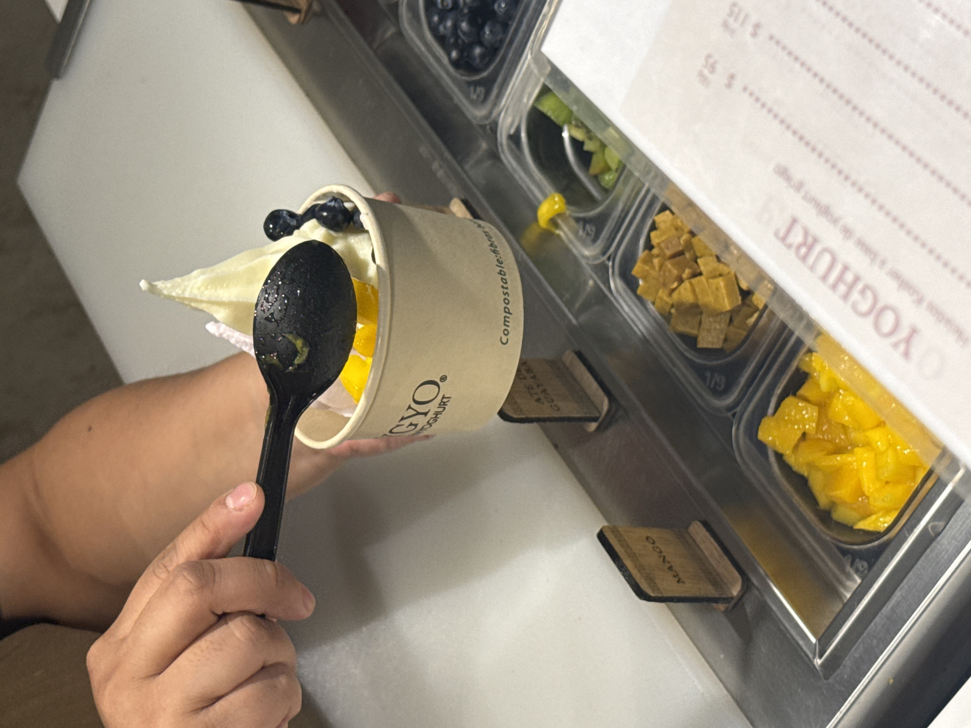 A person is preparing a frozen yogurt dessert at a shop called GYO in Mexico City, Mexico. The person is holding a cup of frozen yogurt with a variety of toppings including blueberries, kiwi, mango, and another yellow fruit in their left hand and a black spoon in their right hand, apparently adding toppings to the cup. The shop appears to be modern and clean, and has a variety of toppings available to customers. There is a sign in the foreground that lists the prices of the yogurt in pesos.
