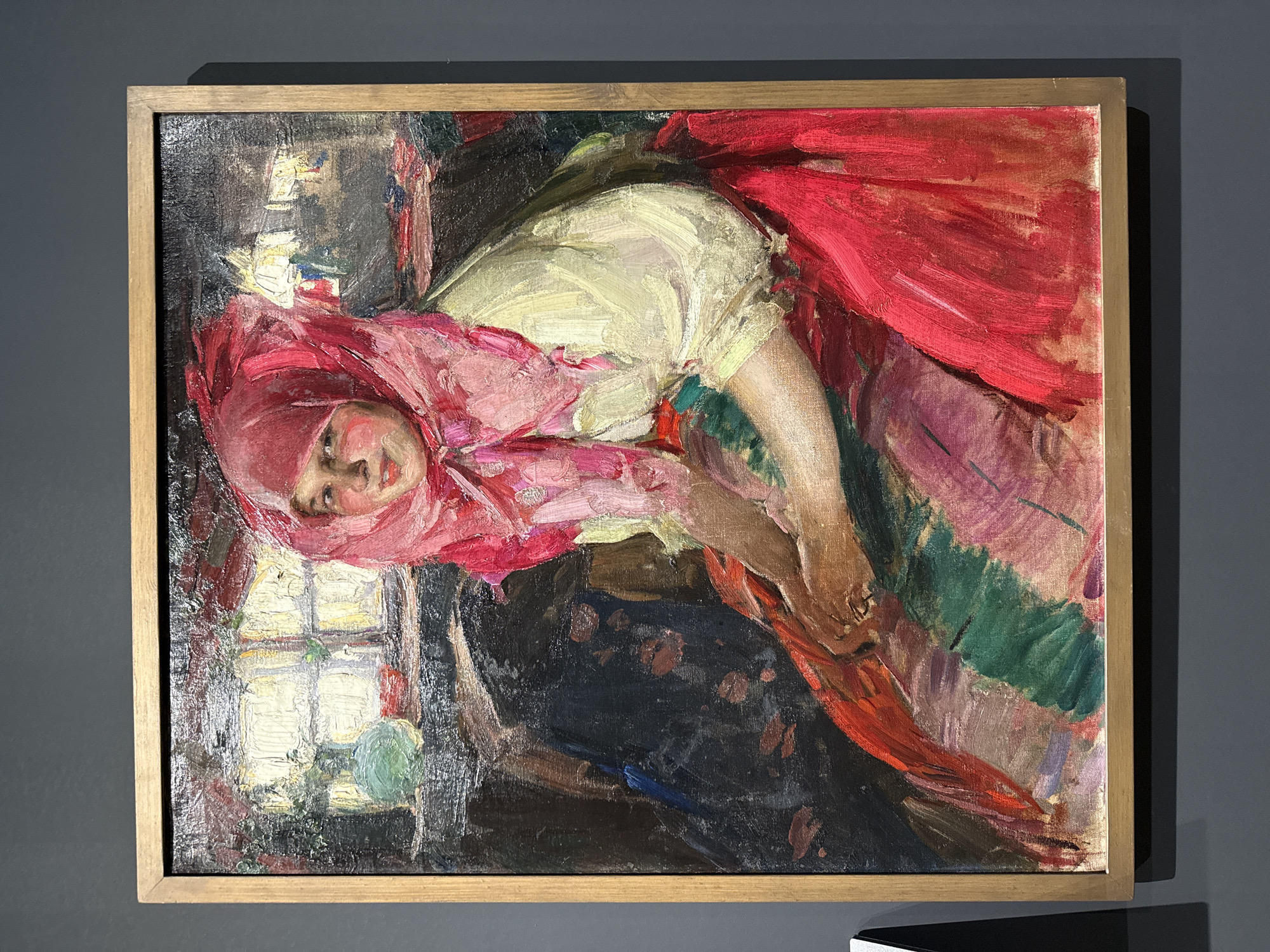A painting depicts a young woman in a vibrant red headscarf and a white blouse. She is seated in what appears to be an indoor setting with a window in the background, suggesting daylight. The artwork is housed in a simple wooden frame and is displayed against a dark gray wall. The painting is located at the Perm State Art Gallery in Perm, Russia.
