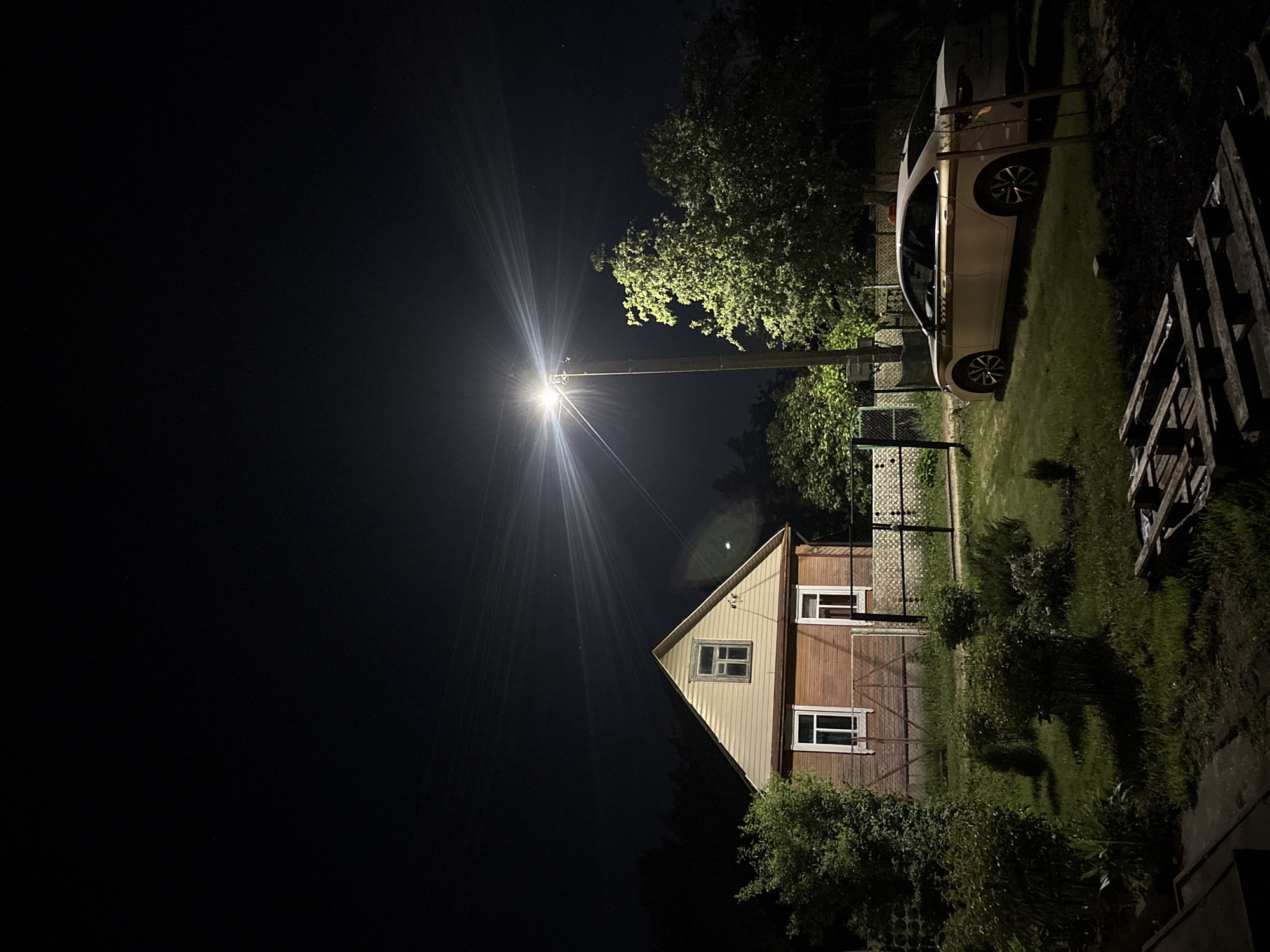 At night in Пярэжырскі сельскі Савет, Belarus, a two-story house is illuminated by a streetlight. The house has wood siding on the lower level and light-colored siding on the upper level. A white car is parked near a fence. The lawn is overgrown. A wooden structure, possibly pallets, sits on the right edge of the frame. The sky is dark.