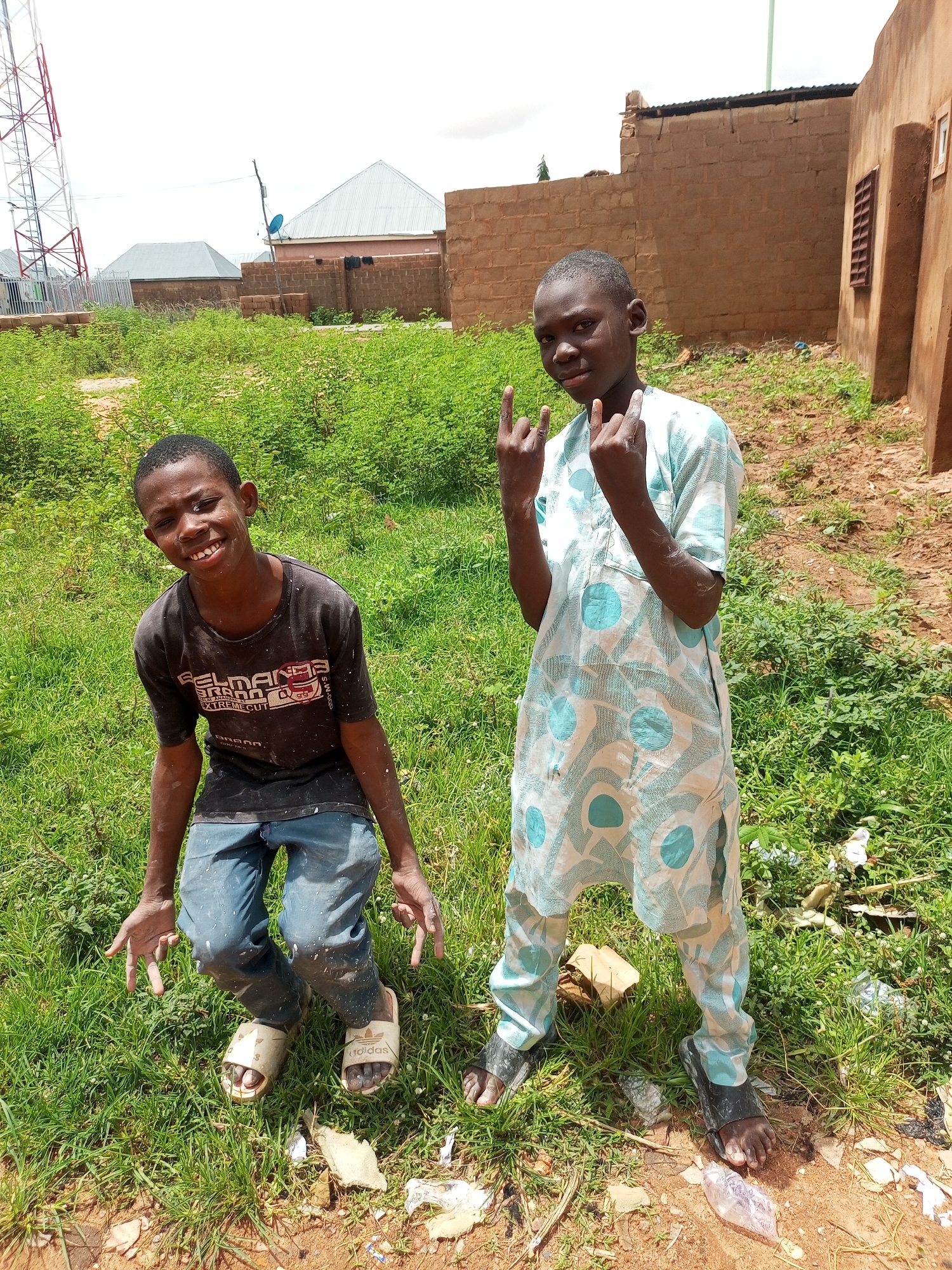 Two young boys pose for a photo in an outdoor, rural setting. The boy on the left, wearing a black t-shirt and blue jeans, is crouching with his hands outstretched. The boy on the right, dressed in a light blue and white patterned traditional garment, stands with his hands raised, making a "rock on" gesture with both hands. The background consists of overgrown green grass, some scattered debris, and rustic brick buildings with corrugated metal roofs. A large radio tower is visible in the distance. The scene appears to be a casual, everyday moment in a Nigerian village.