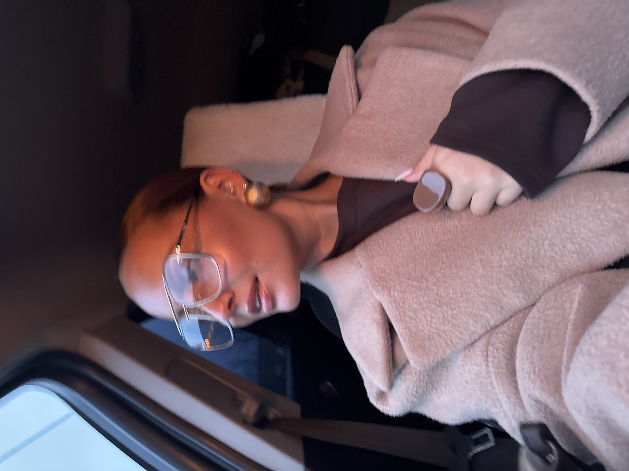 A person is seated in what appears to be the passenger seat of a vehicle. They are wearing glasses, a dark-colored shirt, and a light-colored fuzzy jacket. The person is holding onto the seatbelt or a similar object. The background suggests an interior of a vehicle with a window visible. The image does not contain enough information to place the location in Chaikovsky, Russia.