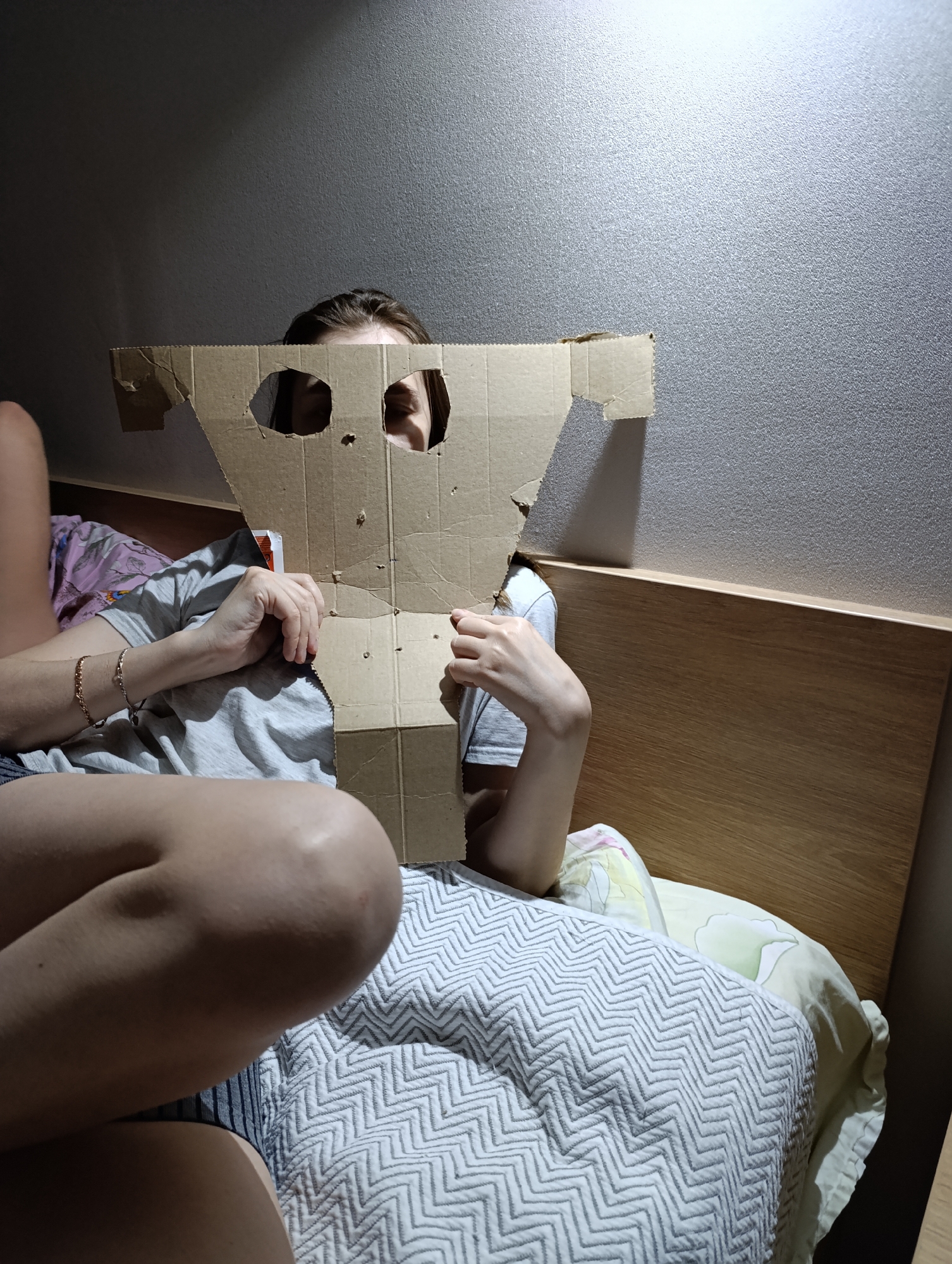 A person lies on a bed in a room in Chaikovsky, Russia. The person holds a crudely-cut cardboard mask in front of their face, obscuring most features except for the eyes visible through cut-out holes. The room is dimly lit. A headboard is visible behind the person. A patterned blanket covers the bed.