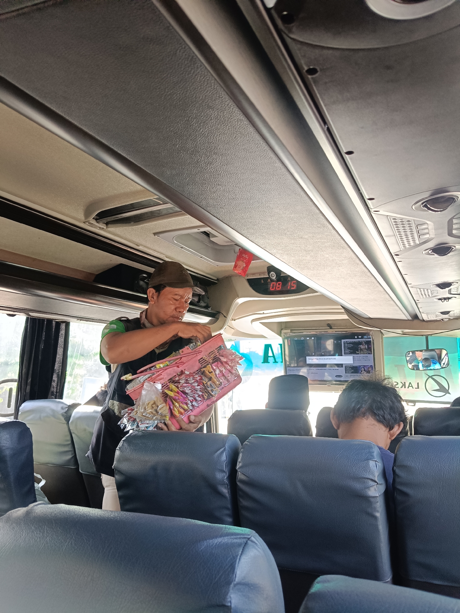 A man wearing a dark vest over a green shirt and a brown cap holds a pink plastic basket filled with individually wrapped snacks, such as candy and cookies. He is standing in the aisle of a bus, looking down at the basket and appears to be arranging or distributing its contents. The bus interior is visible, with rows of dark blue seats. A digital clock on the ceiling displays "08:15". A screen above the seats shows a video with text in Indonesian and a mirrored reflection of the bus interior. The scene appears to be within the city of Kedungrejo, Indonesia.