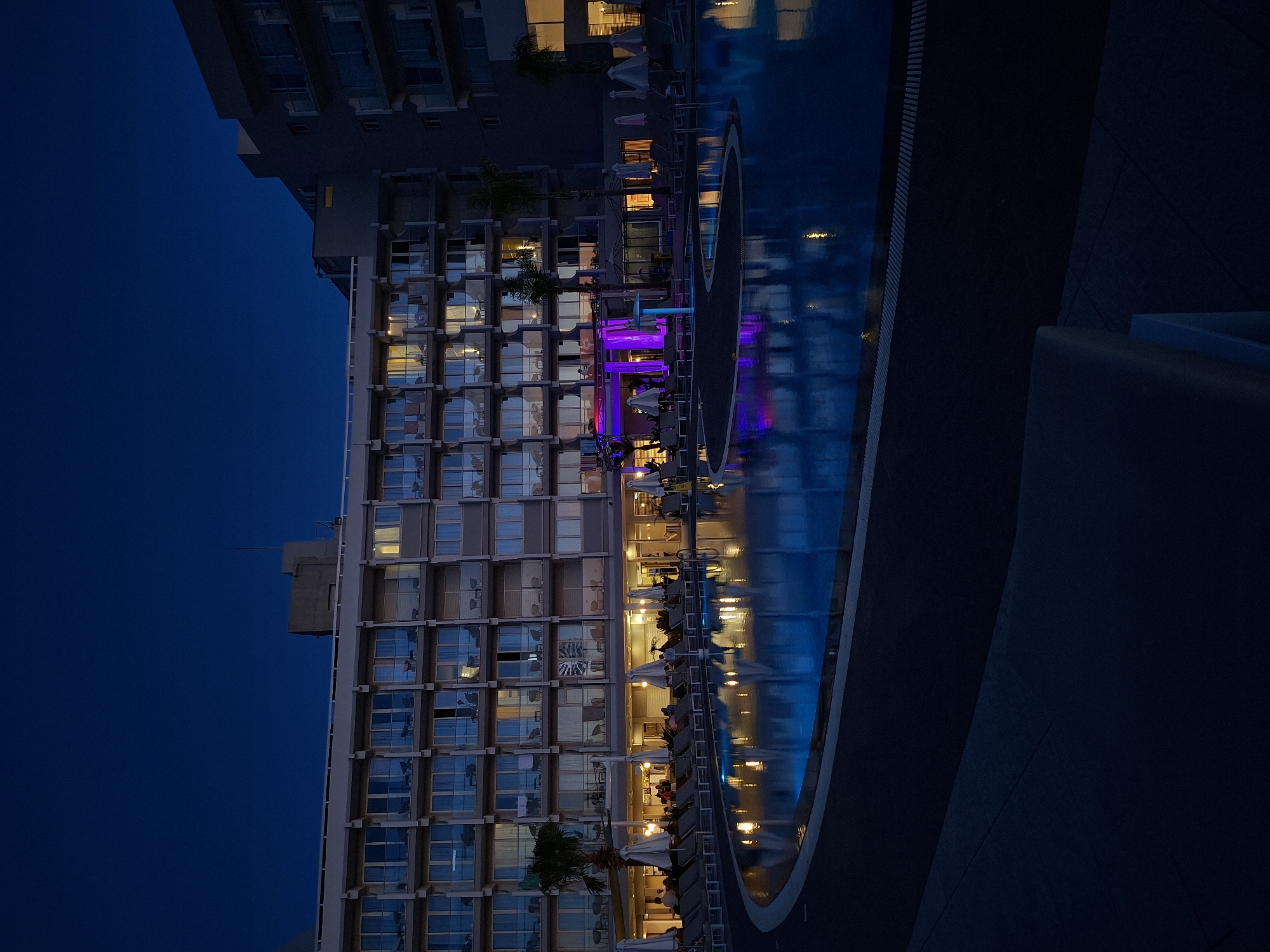 The image depicts a hotel in Protaras, Cyprus at dusk. The hotel building features multiple floors with many windows reflecting the light, casting a grid-like pattern on the facade. A swimming pool runs alongside the hotel with reflections of the hotel lights and lounge area. Lounge chairs with white umbrellas are visible next to the pool, with several people seated. The sky is a deep blue, and the light level suggests it is early evening.