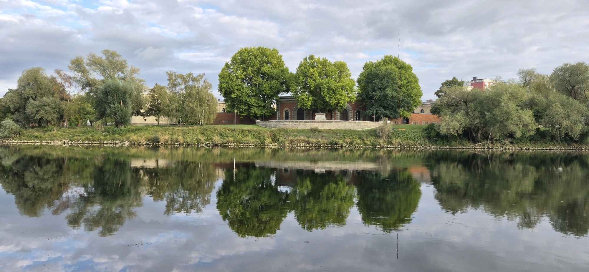 This is a wide, horizontal, eye-level, outdoor, landscape photograph of the Donauregion in Ingolstadt, Germany, taken on a slightly overcast day. The image captures a tranquil scene along the Danube River, with lush greenery on the riverbank and a reflection of the sky and trees on the water's surface.
In the foreground, the dark, reflective surface of the Danube River dominates the lower half of the frame. The water mirrors the sky, clouds, and the trees on the opposite bank with remarkable clarity. The sky is a mix of pale blue and white clouds, with the clouds casting their reflections on the water.
The middle ground features the verdant riverbank, lined with a variety of trees. Prominent among them are several large, mature trees with full, green canopies, some of which appear to be sycamores. To the left, a cluster of lighter-colored trees, possibly birch, stands out. Behind the trees, there are buildings constructed of brick, appearing to be part of a historical complex, possibly a former fortress or military installation, given Ingolstadt's history. The buildings have arched windows and doorways. A thin, dark, vertical pole extends upwards from the grassy area behind the trees, near the center of the frame.
The background consists of more trees and hints of urban structures further away, including a building with a red roof. The overall impression is one of natural beauty harmoniously coexisting with historical architecture. There are no people visible in the photograph. The lighting suggests it is daytime, possibly late afternoon or early morning, judging by the soft, diffused light.