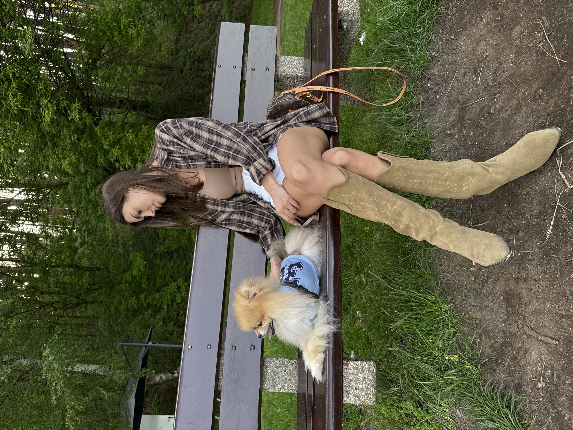 A young woman is sitting on a park bench in an outdoor environment. She is wearing a brown plaid shirt, a tan undershirt, white shorts, and high suede boots. To her right sits a small, fluffy Pomeranian dog wearing a blue sports jersey with the number 3 on it. The woman appears to be interacting with the dog, possibly petting it. A brown purse sits on the bench to the woman's left. The park has green grass, trees in the background, and a dirt and grass area where one of the woman's boots rests.