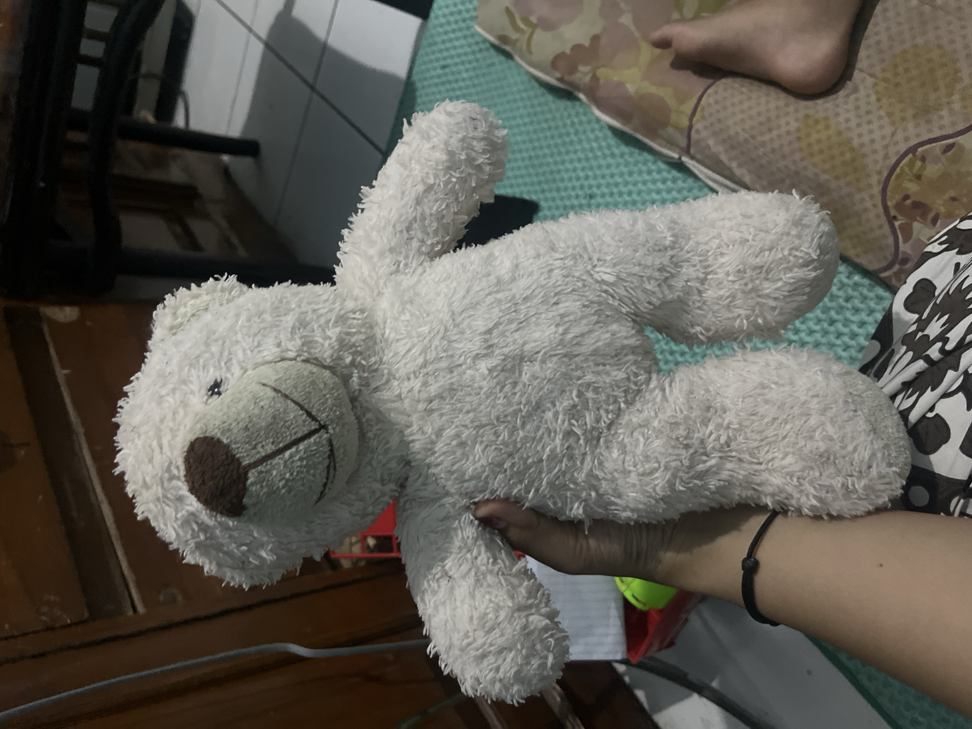 A close-up shot shows a white teddy bear being held in someone's hand. The teddy bear has a brown nose and what appears to be stitched-on eyes. The arm holding the teddy bear has a simple black bracelet. The background includes a wooden floor, a bed with a green cover, and a pillow with a pattern. The picture appears to have been taken indoors, likely in a domestic setting. The location is Kranggan, Indonesia.