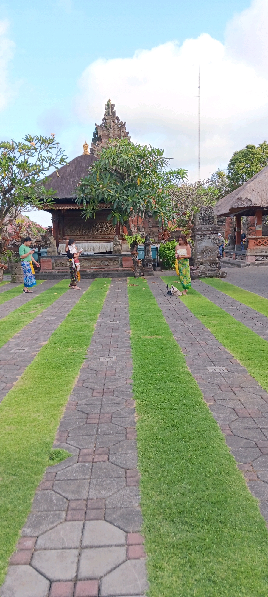 The image depicts a paved walkway with alternating strips of grass, leading towards a temple complex in Sukawati, Indonesia. The temple features traditional Balinese architecture with a tiered roof and intricate carvings. Three individuals are visible in the scene. On the left, a person wearing a green shirt and a sarong walks on the path. In the center, another person holding a child is standing near the temple entrance. To the right, a person in a green and yellow sarong is looking at their phone. The sky above is partly cloudy, and trees surround the temple grounds. A tall tower, possibly a communication mast, is visible in the background.