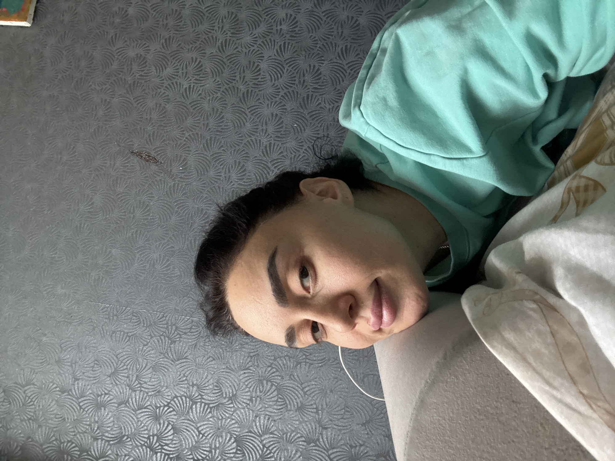 An indoor image captures a person, likely female, lying down and looking directly towards the viewer with a relaxed and subtly smiling expression. The individual has dark hair, prominent dark eyebrows, and full lips. They are dressed in a light teal or mint green long-sleeved top, possibly a hoodie, and their head is resting on a soft, light grey or off-white surface, which could be a pillow or part of a sofa or bed. A thin white wire, possibly an earbud cord, is visible near their chin.
The background features a dark grey wall covered in a textured wallpaper or decorative panel. The pattern on the wall consists of repetitive, fan-like or scalloped motifs. A faint horizontal seam runs across this patterned surface. To the right side of the frame, a white and beige patterned fabric, resembling a blanket or bed linen, is visible, adding a warm tone to the composition. In the upper left corner of the patterned wall, a small, dark, elongated piece of debris or a small insect can be observed. The lighting suggests it is daytime. The scene is set indoors, and the provided location context is Tbilisi, Georgia. There is no visible text in the image.