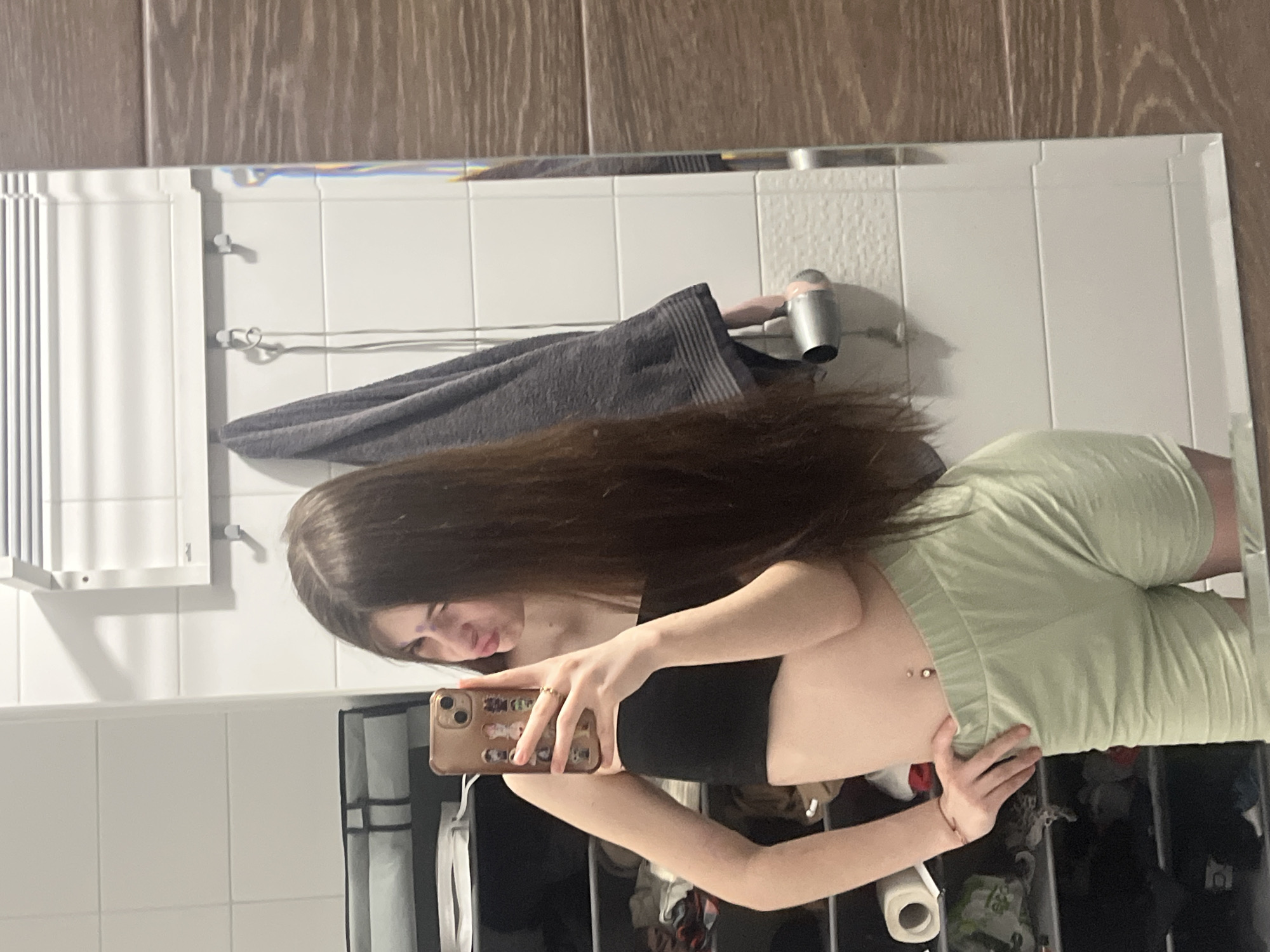 The image displays a mirror selfie taken by a young woman in what appears to be a bathroom or utility room, likely in Saint Petersburg, Russia. The woman, with long, straight, dark brown hair, is wearing a black strapless crop top and light green elastic-waist shorts. A belly button piercing is visible on her midriff. She holds a gold-colored smartphone with a decorated case, capturing her reflection in a large mirror. Her expression is a soft, relaxed smile or pout.
The setting, reflected in the mirror, features white rectangular wall tiles, topped by darker wooden paneling. To the left of the woman, a white radiator or heating unit is mounted on the wall. Behind her, a dark gray towel hangs on a hook next to a silver hair dryer. The lower part of the reflection reveals what seems to be a storage area or shelving unit containing various items, including a roll of toilet paper, reinforcing the impression of a bathroom. The lighting suggests an indoor, artificial light source. No discernible text is visible on any objects. The original image is rotated 90 degrees counter-clockwise.
