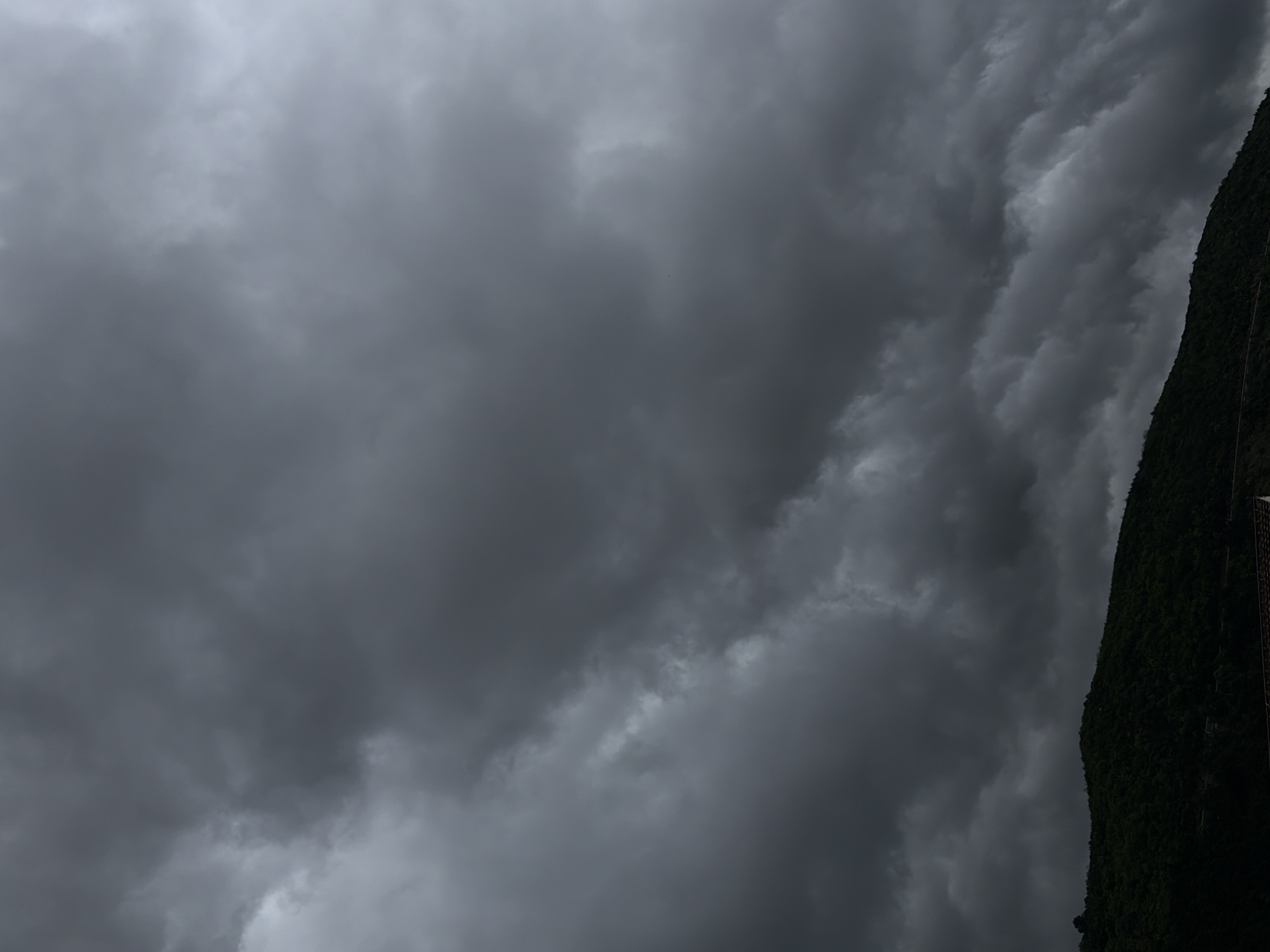 The image captures a dramatic sky filled with heavy, dark grey storm clouds, occupying most of the frame. To the right, a steep, dark green, heavily vegetated hillside or mountain face rises vertically, appearing as a dark silhouette against the turbulent sky. The dense foliage on the slope suggests a natural, possibly rugged, landscape.
The overall atmosphere is moody and overcast, indicating impending rain or stormy weather. The dark tones of the clouds range from deep charcoal to lighter grey, with some areas appearing almost black, creating a sense of depth and movement. The contrast between the dark clouds and the even darker green of the mountain accentuates the dramatic nature of the scene. No specific activities or people are visible, and the scene is entirely natural. There is no visible text in the image. Given the context, this scene is set in Budva, Montenegro, likely showcasing its mountainous coastal environment under a cloudy sky.