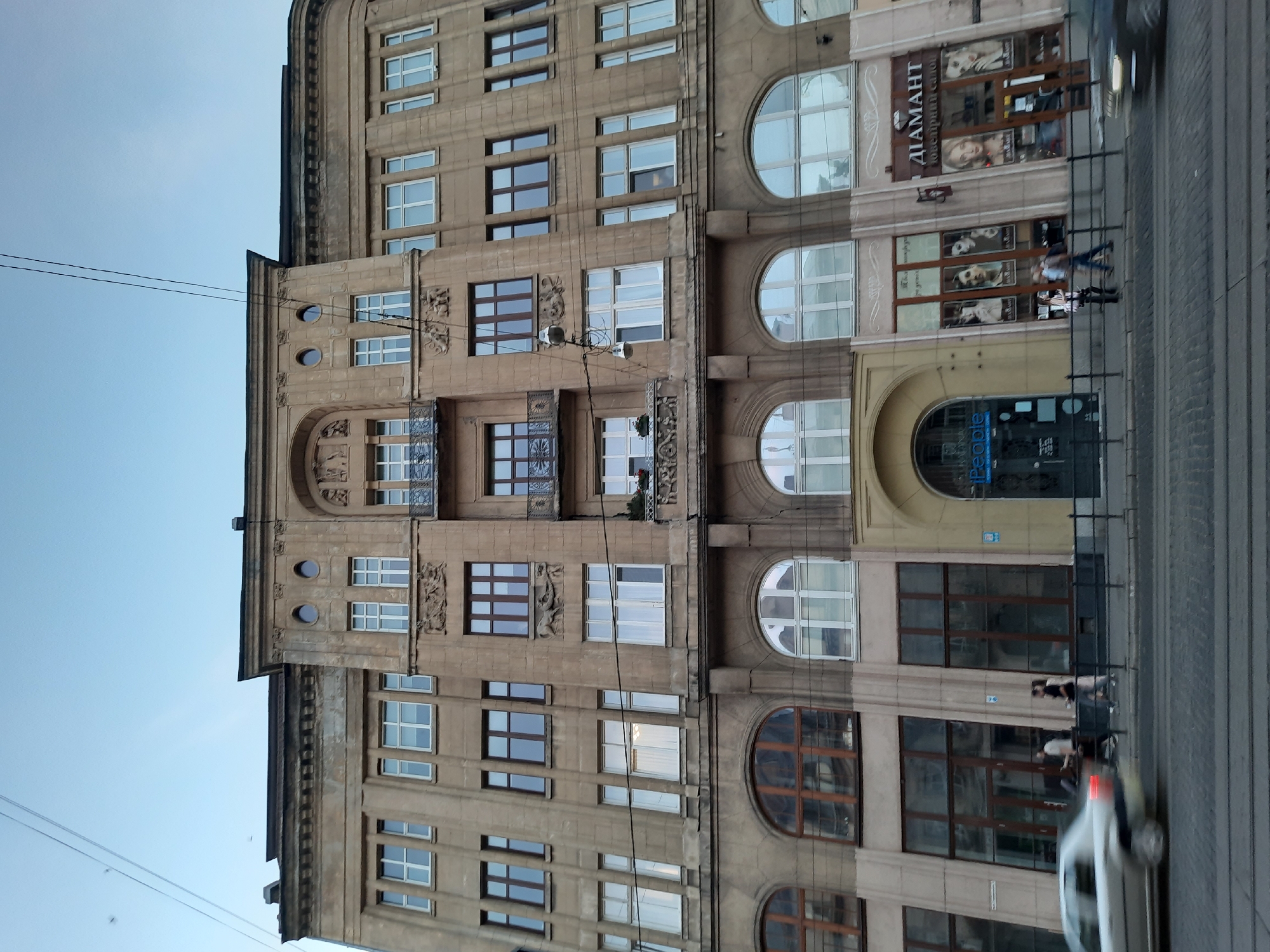 The image shows a wide-angle, low-angle view of an ornate, multi-story building with a light brown facade, located in Lviv, Ukraine. The architecture features decorative elements above the ground floor, including intricate carvings and arched windows. The ground floor is occupied by several storefronts. To the left, there is a storefront with large, dark-framed windows, followed by an arched entrance with a blue "iPeople" sign. To the right of the iPeople entrance, there are two shops with large display windows showcasing portraits of women and text in Cyrillic. The shop on the far right has a prominent sign reading "ДІАМАНТ" (DIAMANT) and "ЮВЕЛІРНИЙ САЛОН" (JEWELRY SALON), indicating a jewelry store.
In the foreground, a street with cobblestone paving is visible, with tram tracks running across it. A silver car is blurred in motion on the left side of the image, suggesting traffic. Several pedestrians are walking on the sidewalk in front of the shops. One individual is seen entering the iPeople store, while others are walking past the shop displays. Power lines crisscross the sky above the building. The sky is clear and pale blue, indicating it might be early morning or late afternoon. The overall scene captures a segment of a busy street in Lviv, highlighting its architectural heritage and commercial activity.