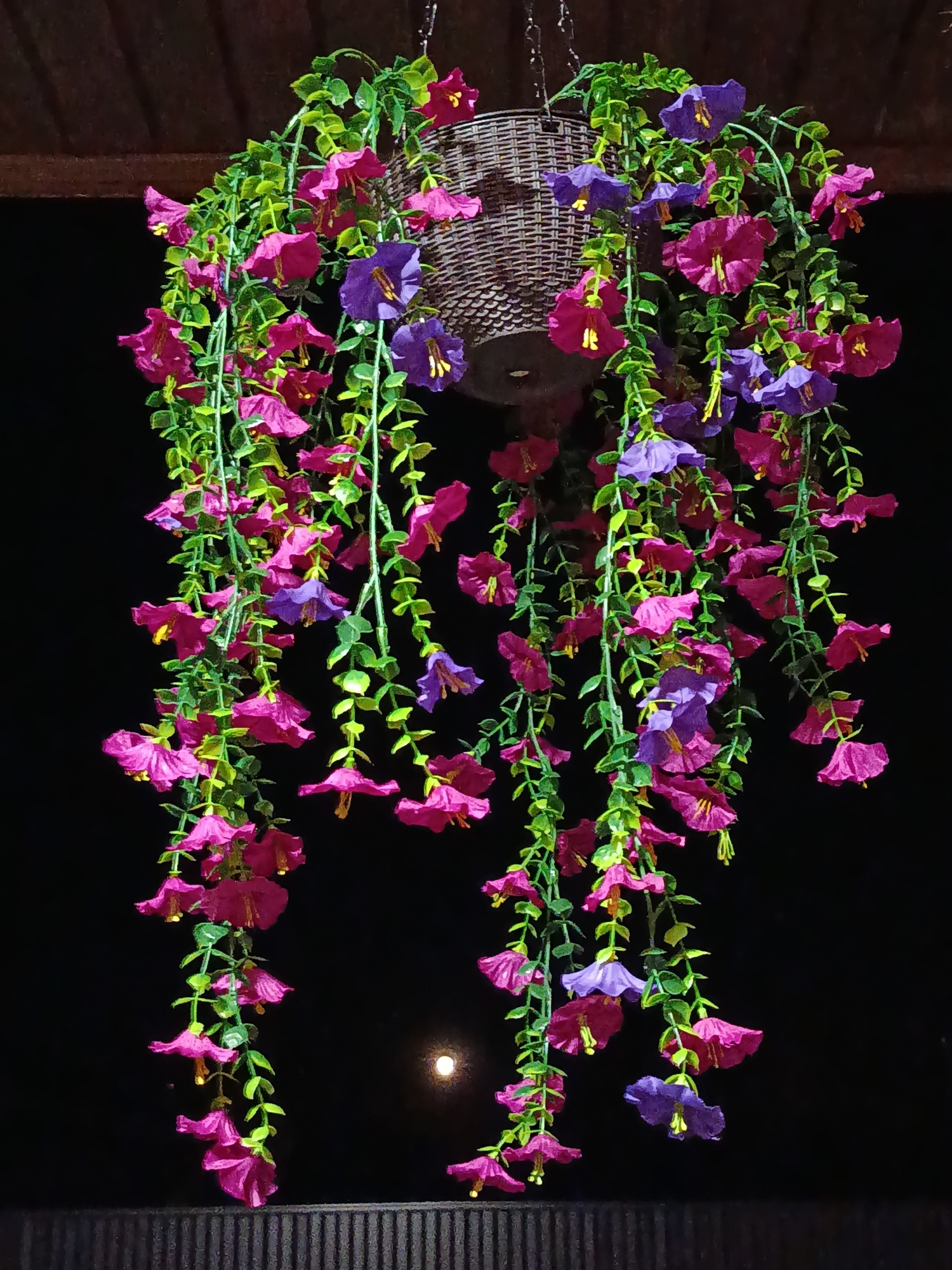 A hanging basket overflowing with artificial pink and purple bell-shaped flowers, suspended from a dark brown wooden overhang. The basket itself is woven and appears to be made of wicker or a similar material. The flowers and their green artificial vines cascade downwards against a black background, creating a stark contrast. In the lower portion of the image, a blurred circle of light, possibly a distant moon or a light source, is visible. Below the hanging arrangement, a textured dark surface, perhaps a fence or railing, is partially visible. This scene is located outdoors in Verea, Russia.