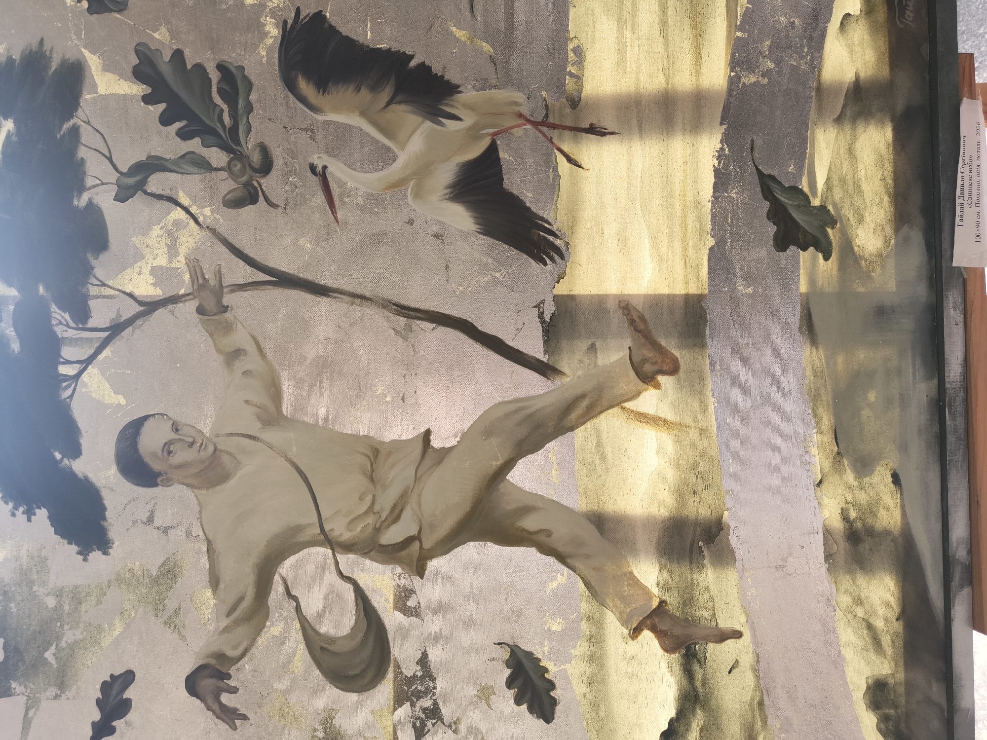 The image is a painting depicting a young man in simple, light-colored clothing, possibly a tunic and trousers, in motion. He has his arms outstretched, his left hand reaching forward, and his right hand slightly back. He appears to be running or leaping across a textured, somewhat abstract landscape. A dark bag or pouch is slung over his left shoulder, trailing behind him.
To his right, a white stork with black wingtips and red legs is also depicted in motion, its wings partially spread as if in flight or landing. Oak leaves and acorns are visible in the upper right portion of the painting, suggesting an autumnal or natural setting. The background is a mix of muted colors and gold leaf, creating a textured and somewhat ethereal effect. The ground beneath the figures is rendered in shades of yellow and grey, with prominent shadows cast by the man and the stork.
A small white label is visible at the bottom of the painting. Although the text is partially obscured and difficult to read entirely, it appears to be a caption for the artwork. The legible parts suggest it might be a title, artist's name, and dimensions, possibly indicating it is an exhibit in Kyiv, Ukraine, as the artist's name "Гайдай Данило Сергійович" and the title "«Свищеве небо»" appear in Cyrillic.