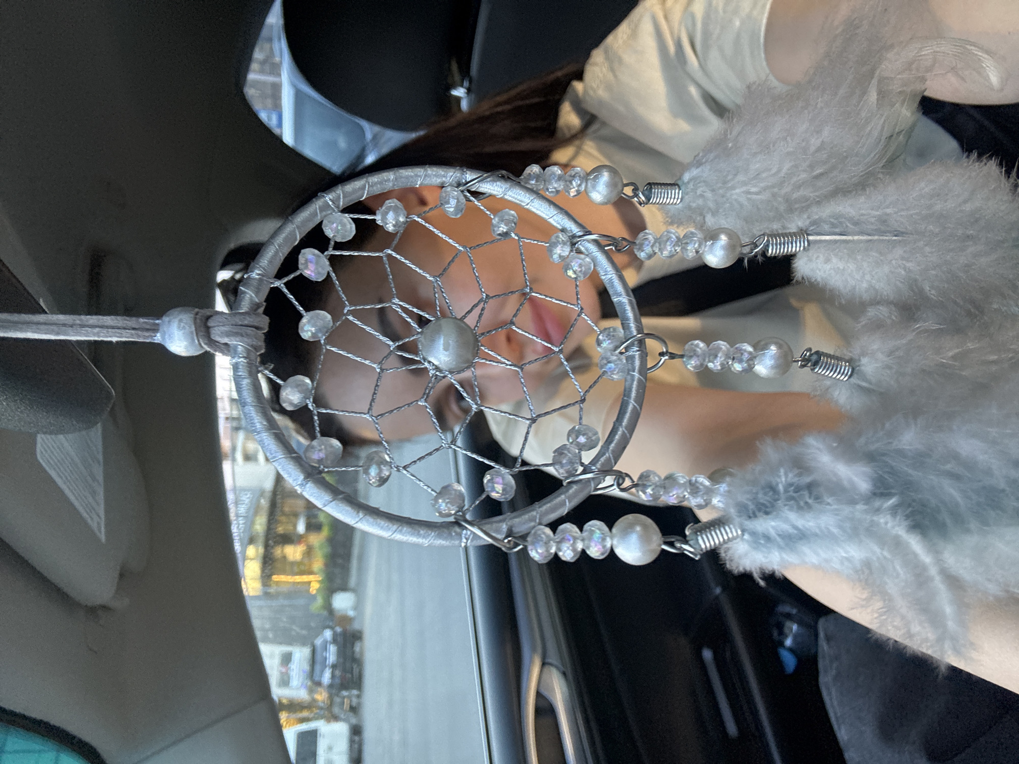 The image depicts a silver-colored dreamcatcher hanging inside a car, with a person's face blurred in the background, visible through the dreamcatcher's web. The photograph is oriented horizontally but described as if upright.
The dreamcatcher, the main subject in the foreground, consists of a silver-wrapped hoop with an intricate web woven from silver thread, adorned with several clear, iridescent beads. Hanging from the bottom of the hoop are multiple strands featuring a sequence of smaller clear beads and larger, pearl-like beads, each terminating in soft, fluffy grey feathers.
Behind the dreamcatcher, a person (likely a woman) is partially seen and out of focus. Her facial features, including parts of her eyes, nose, and mouth, are visible through the dreamcatcher's open patterns. Her tongue is slightly visible, suggesting a relaxed or pensive expression. She has long, dark hair and is wearing a light-colored top.
The setting is inside a car, indicated by elements such as the car's ceiling, a sun visor, and part of a door panel visible around the dreamcatcher. Through the car windows, an out-of-focus view of an urban or suburban landscape can be seen, with buildings and what appears to be a street scene. The lighting suggests it is daytime, possibly late afternoon or early evening, with some distant artificial lights already illuminated. No specific weather conditions are evident. The location context is Tbilisi, Georgia. There is no discernible text visible in the image.