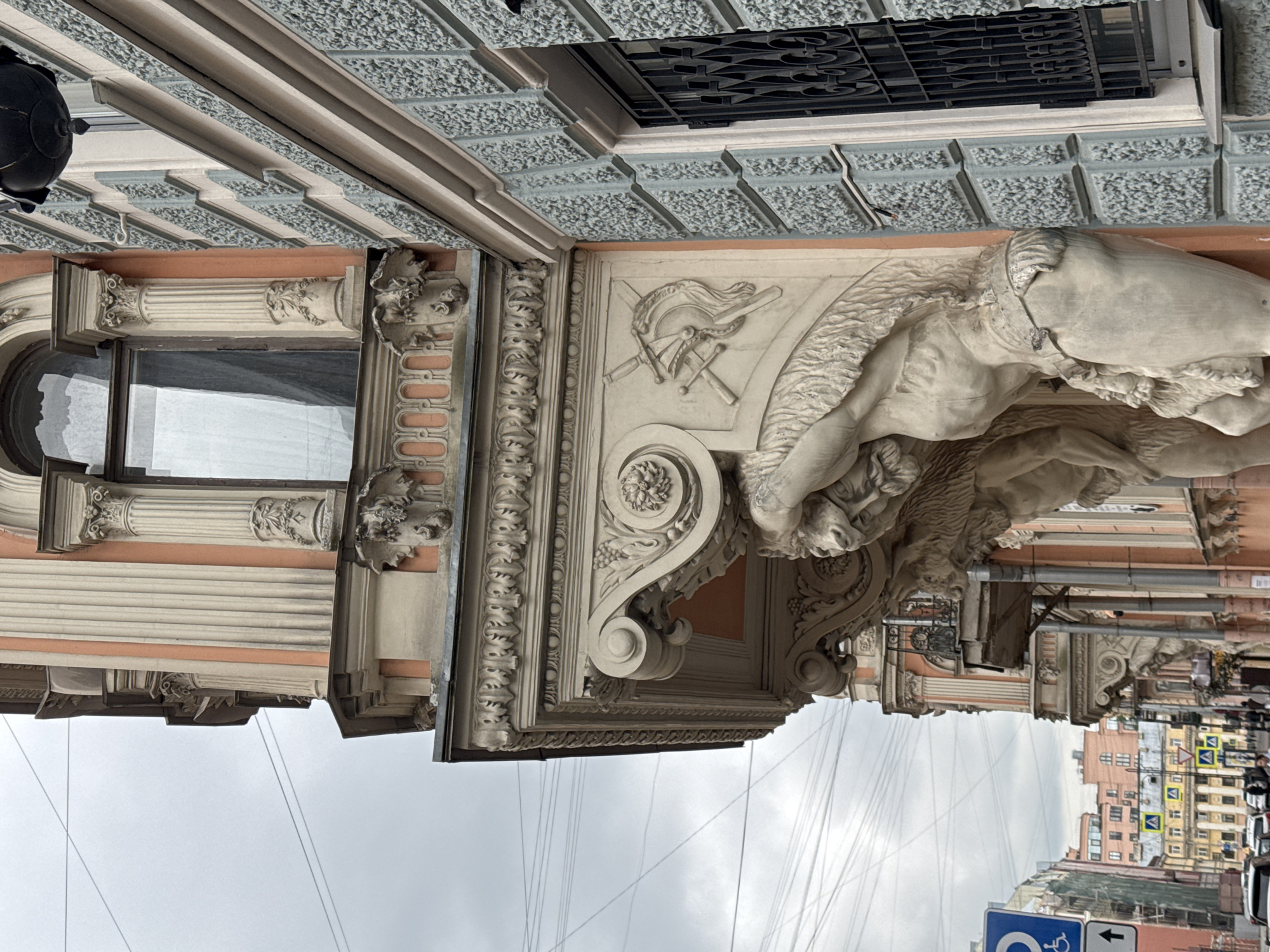 A street-level, medium-angle shot of a corner of a building in Saint Petersburg, Russia, taken on an overcast day. The primary subject is the building's architectural detail, specifically a sculptural element depicting a figure, possibly a lion or griffin, supporting a portion of the building. The sculpture is light-colored and intricately carved. Above the sculpture, there is a frieze with crossed swords and a helmet or torch emblem. A window with ornate stone frame sits to the left of the sculpture. The building's facade features a combination of colors including a light peach or pink, light gray, and darker gray. Above the facade, a dark grille can be seen, likely covering a window or opening. In the lower right corner, there are glimpses of other buildings in the street, as well as street signs, and pedestrians. Power lines run diagonally across the sky.