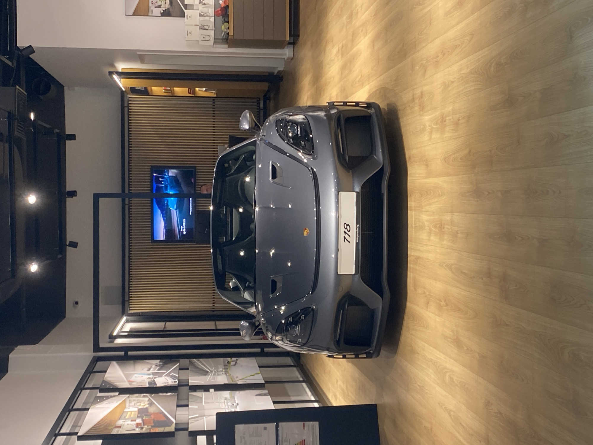 The image shows the front view of a grey Porsche 718 sports car displayed indoors, likely in a showroom. The car is positioned on a light-colored wooden floor. Behind the car, there is a wall with horizontal wooden slats and a television screen displaying an image with blue hues and some text, potentially showing vehicle information. Above the TV, a thin strip of light illuminates the slatted wall. To the left of the TV, a person is partially visible, looking towards the screen.
The foreground features part of a display stand with several framed images showing cars and interior design elements. The overall setting appears to be a modern and well-lit commercial space, possibly a car dealership. The lighting suggests it's daytime, but the indoor setting makes it difficult to determine the exact time or weather conditions outside. The focus is on the car, highlighting its design with sleek lines and prominent headlights. The license plate area is visible, with a white plate displaying the number "718" and some smaller text, possibly a website address, which is not clearly legible. The car has a sporty and luxurious aesthetic, typical of high-end automobile showrooms. The backdrop suggests a clean and organized environment designed to showcase the vehicle.