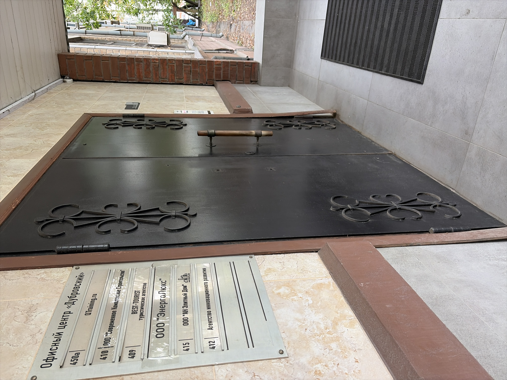 The image shows the entrance to an office center with a sign on the left and a large dark metal door on the right. The sign, titled "Офисный центр «Дубровский»" which translates to "Office Center 'Dubrovsky'", lists several companies with their office numbers. Among them are "QTraining.ru" at office 450a, "ООО «Современное Комплексное Строительство»" at office 410, "BEST-TOURIST туристическое агентство" at office 409, "ООО "ЭнергоЛюкс"" at office 415, and "Агентство инновационного развития" at office 412. The door has decorative wrought iron elements. To the right of the door, there is a brick wall, an air conditioning unit mounted on the wall, and a doormat on the tiled floor. The overall environment suggests an urban setting, likely an office building entrance in Moscow, Russia, given the Cyrillic script on the signage. The time of day appears to be daytime with natural light illuminating the scene. There are no people visible in the image.