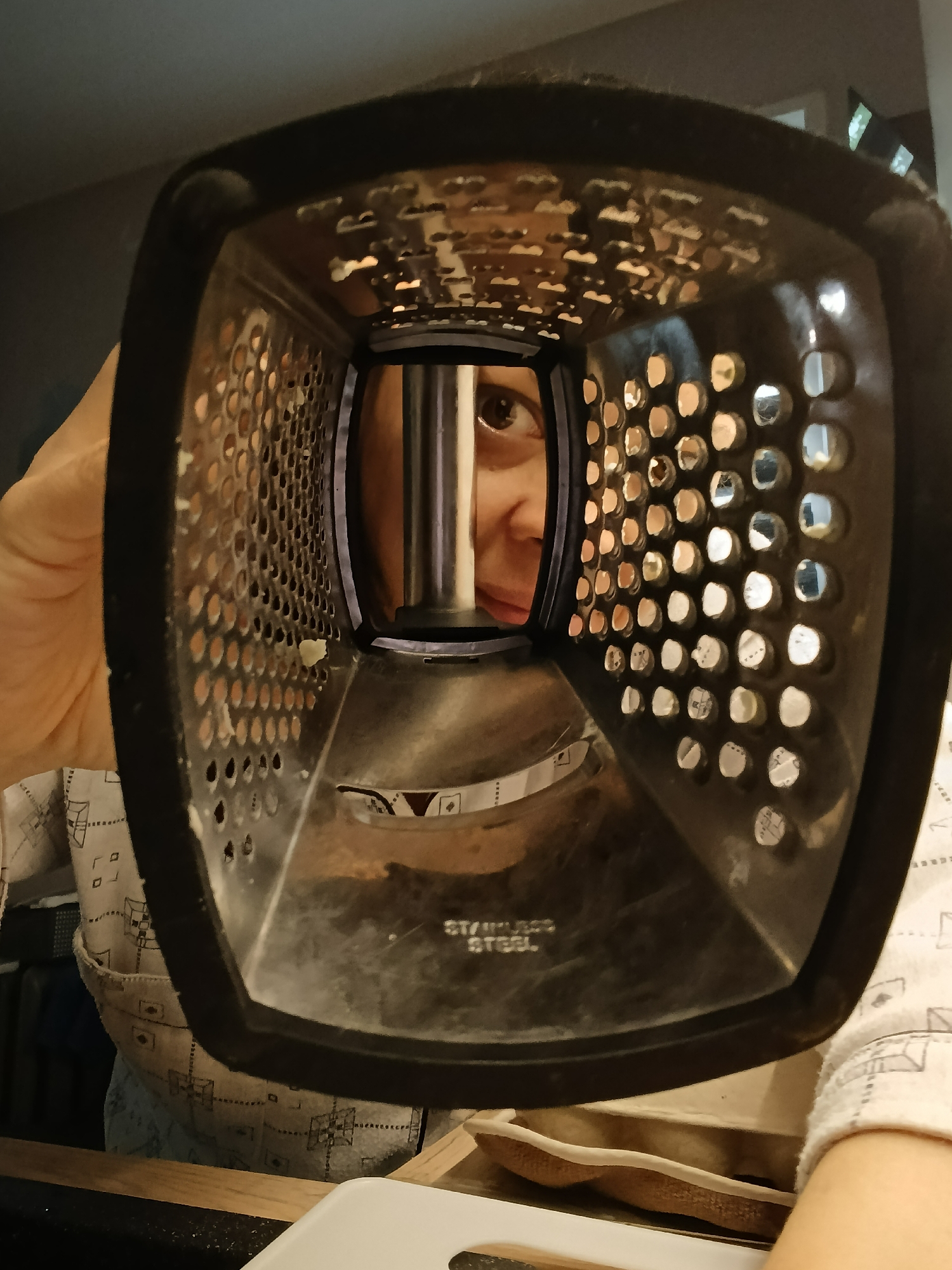 The image is a close-up, POV shot looking through the openings of a metal grater. A person's eye and part of their face are visible through the center of the grater, with their hand holding the grater. The grater has holes of varying shapes and sizes, and some remnants of what appears to be grated food are visible on its surface. The background is blurred but suggests an indoor setting, possibly a kitchen, with hints of clothing and a countertop. The image does not contain any recognizable landmarks or details that would indicate its location in Saratov, Russia, or any specific city.