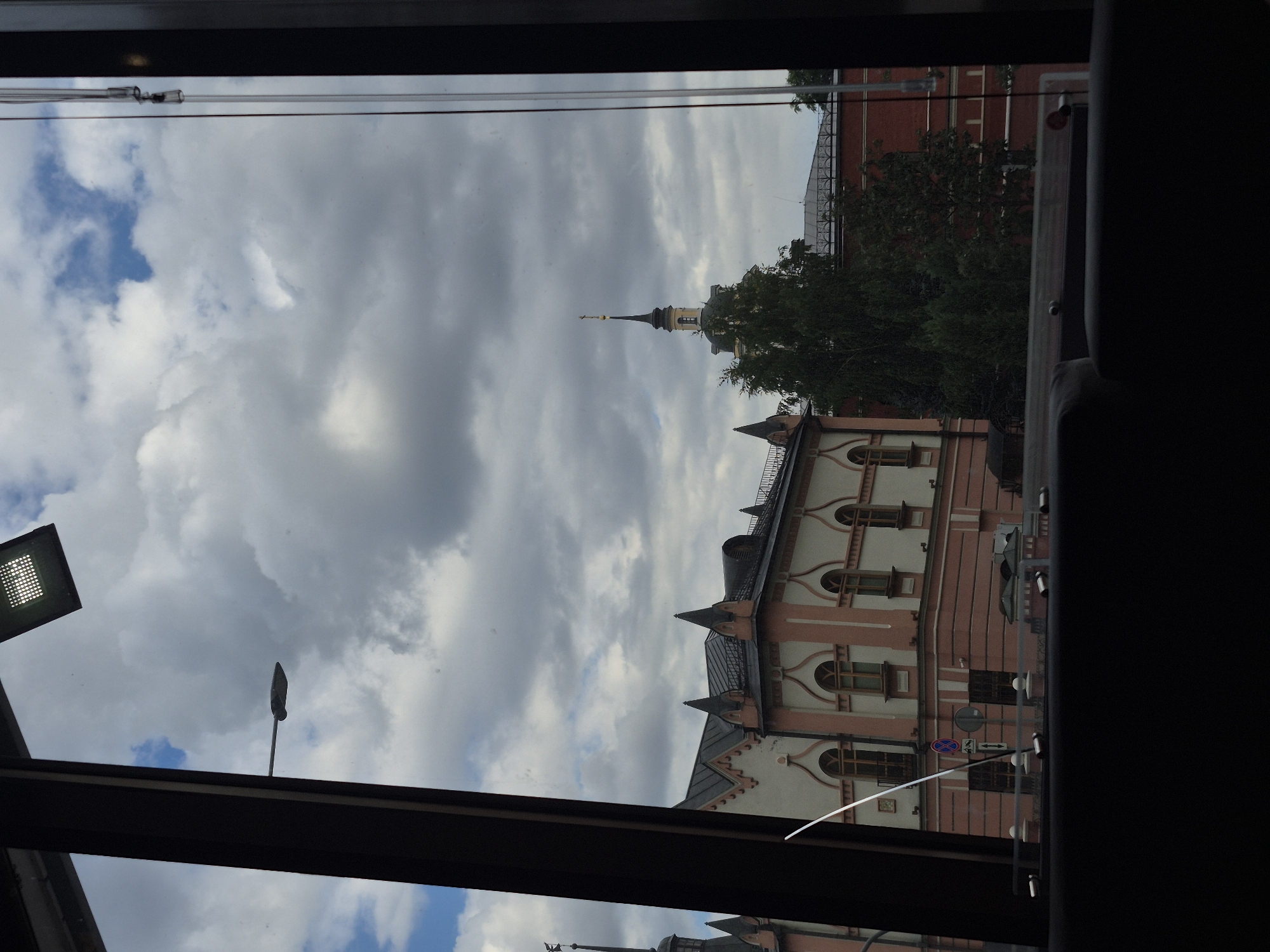 The image, captured through a window, shows a cloudy sky over buildings in Moscow, Russia. In the foreground, a dark, modern window frame and a dark interior seating area are visible. Beyond the window, a building with pink and cream-colored facades and pointed roof details stands out. To the right of this building, a large green tree partially obscures a red brick building. A distinct spire with a cross tops a structure further in the background. A street lamp is visible on a pole, and a modern, rectangular light fixture is mounted above the window frame. The scene suggests an urban environment on an overcast day.