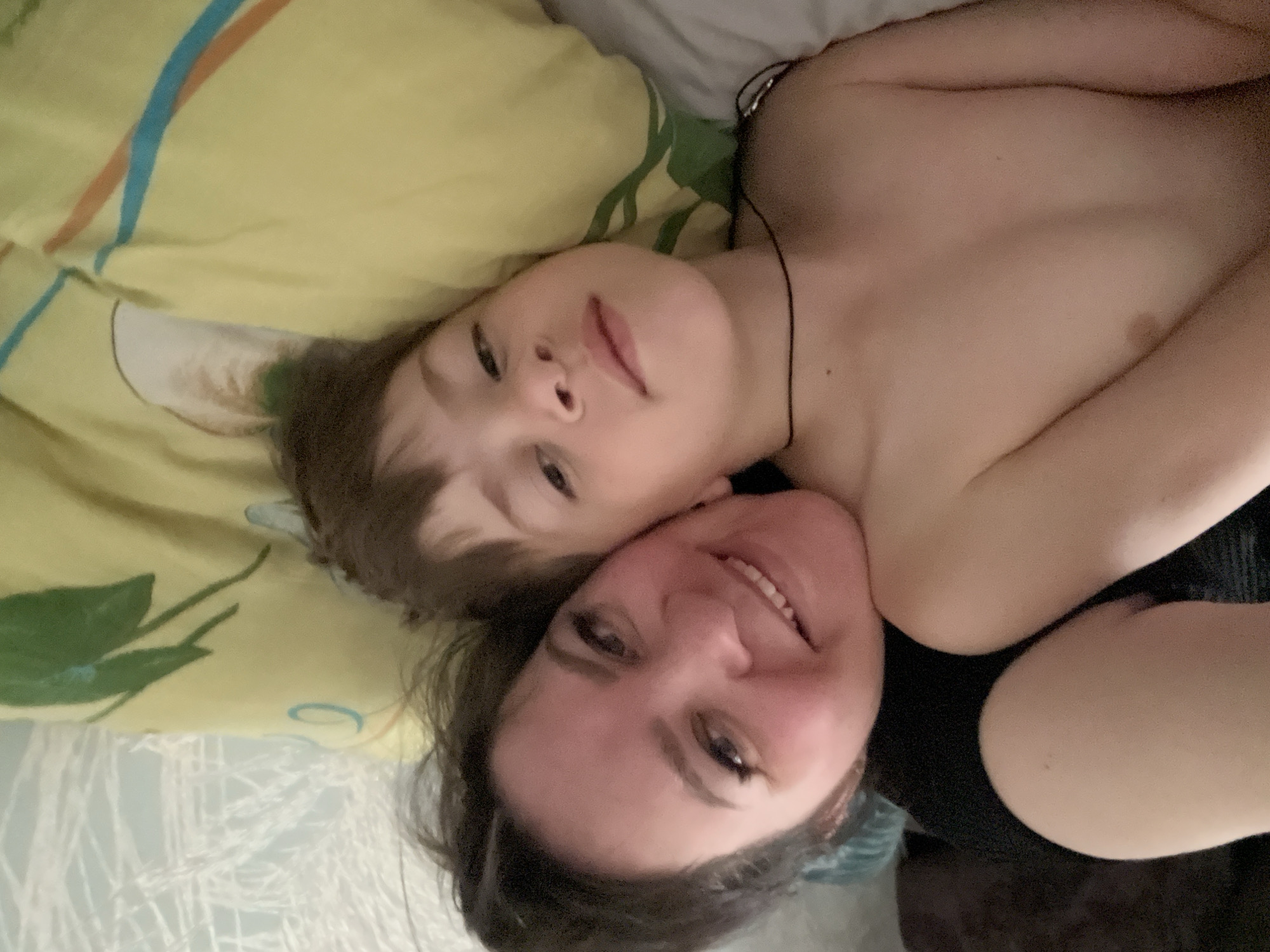 A woman and a young child are lying on a bed. The child is lying on a yellow pillow with a blue and orange stripe, as well as a green leaf print. The child has light hair and eyes. The woman has dark hair and is smiling. The woman is wearing a dark-colored shirt. The background is indistinct. It is not possible to determine the location as Noviy, Russia from the image.