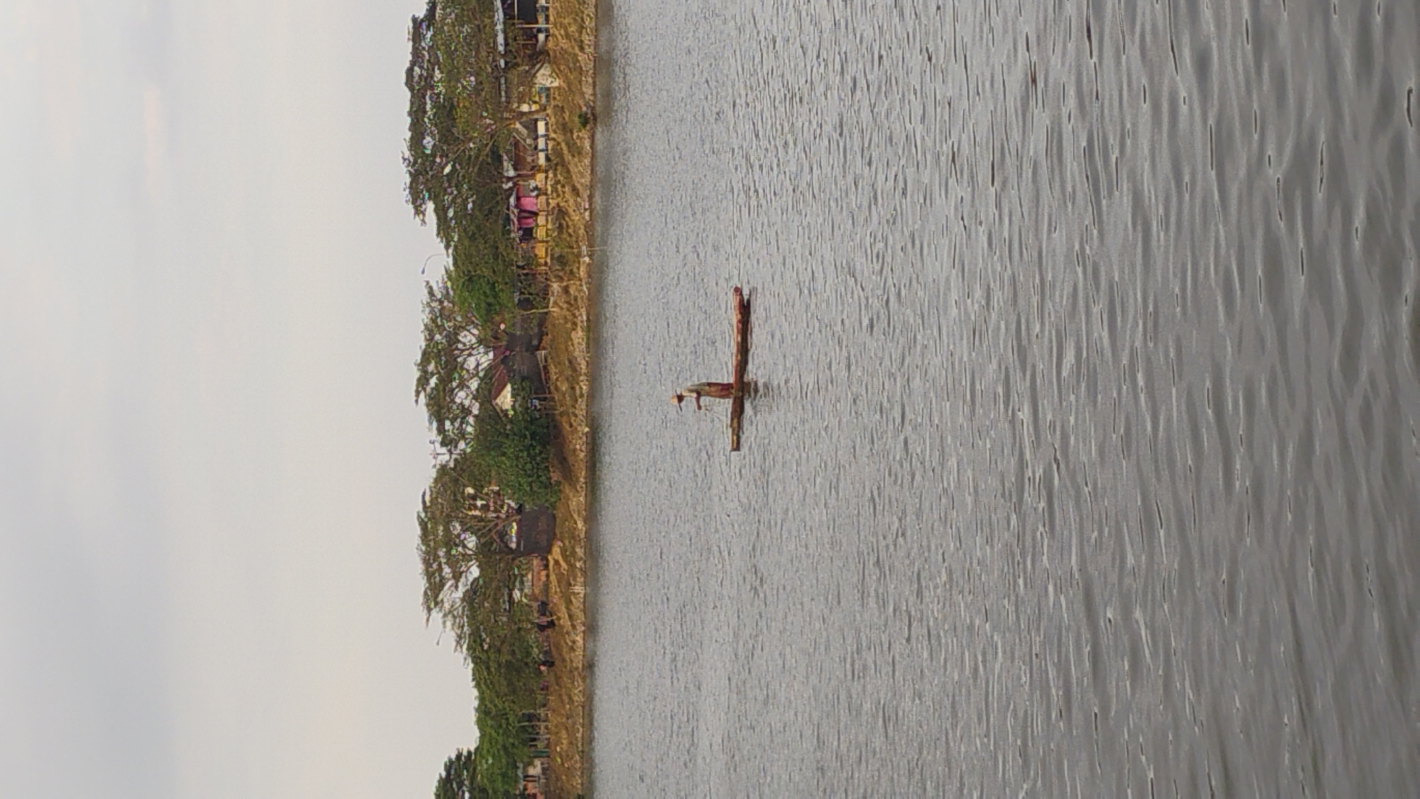 A man stands on a small, narrow wooden raft in the middle of a calm body of water. He is wearing a hat and appears to be fishing, holding a long object in his hands. The raft is a simple, flat structure, possibly propelled by poles. The water has a rippled surface, reflecting the overcast sky. In the background, a tree-lined shore is visible, with several buildings nestled amongst the trees. The area appears to be part of the city of Lhokseumawe, Indonesia, likely a river or a lake within the urban landscape. The overall scene depicts a moment of quiet activity in a semi-rural or suburban setting.