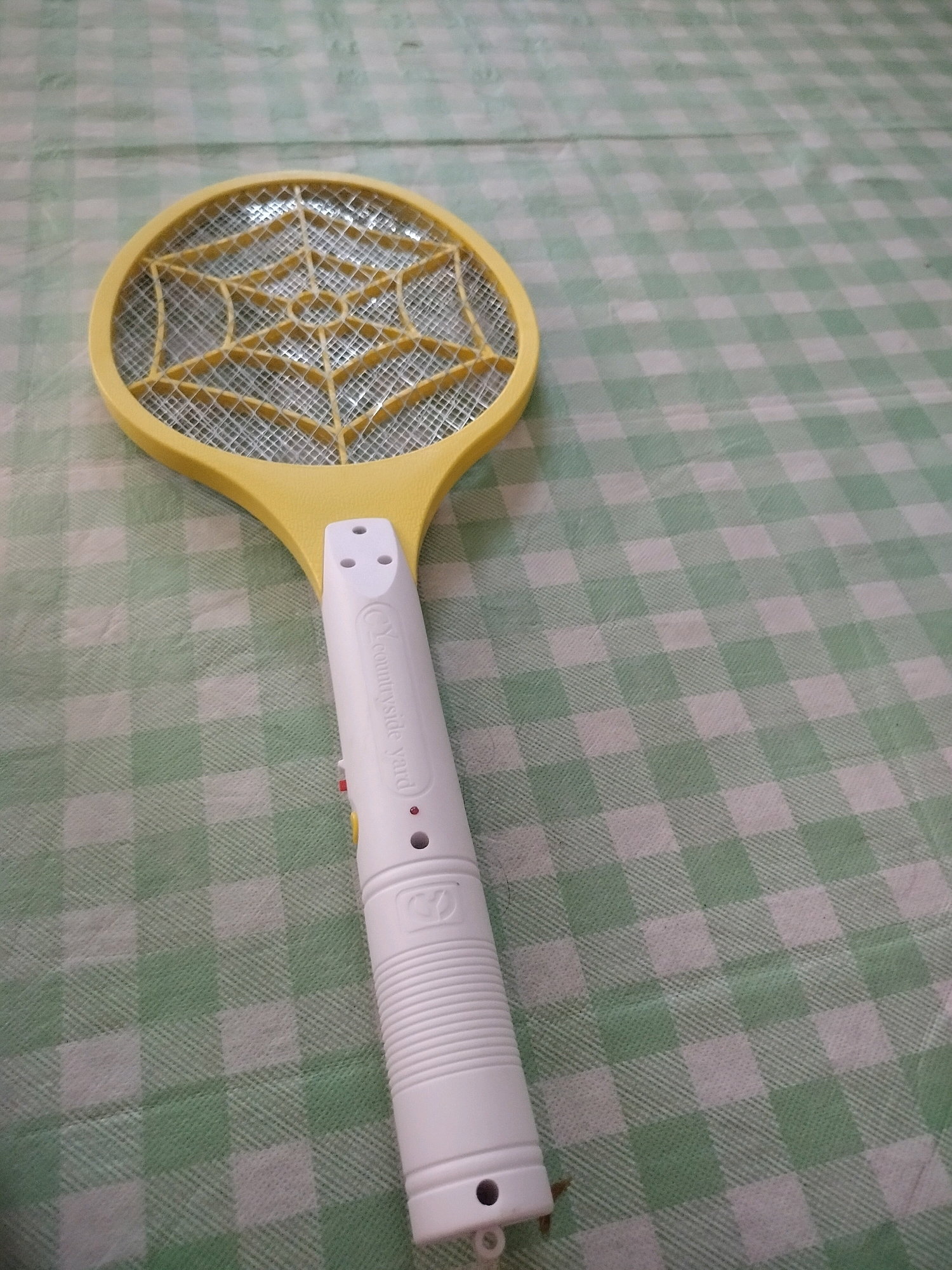 An electric mosquito swatter lies on a green and white checkered tablecloth. The swatter has a yellow frame around the mesh, a white handle with a red button, and a yellow button near the bottom. The words "Y countryside yard" are printed on the handle. There is no indication of any specific location within Kroya, Indonesia.