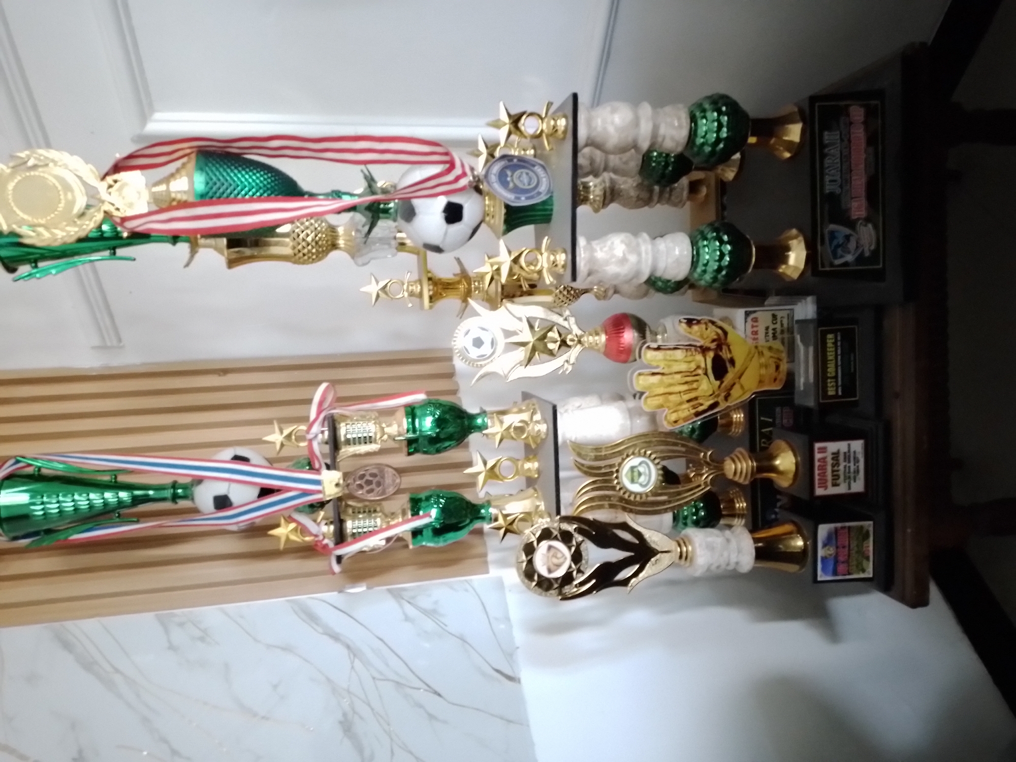 The image displays a collection of trophies and medals displayed on a wooden shelf in what appears to be a domestic setting. The trophies are varied in size and design, featuring predominantly green and gold colors, with some incorporating football elements. Some trophies have ribbons attached, indicating their competitive nature. The background includes a wall with vertical wooden slats and a marbled pattern on the left side. The overall impression is that of a display case for sports awards. The location is identified as Ngancar, Indonesia.