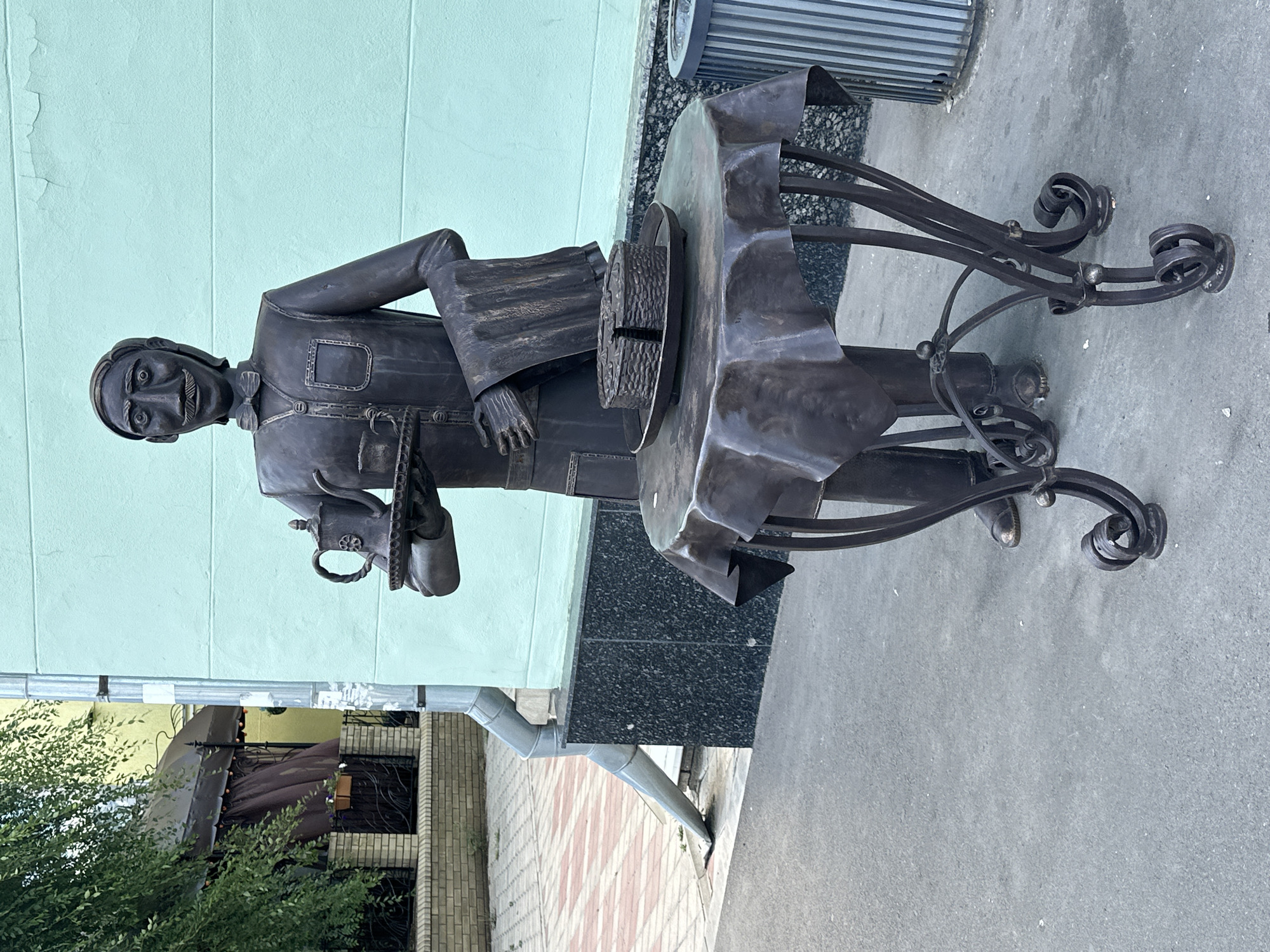 A bronze statue of a waiter stands on a sidewalk in Zhovte, Ukraine, in front of a teal-colored building. The waiter, depicted with a mustache and bow tie, holds a tray with a teapot and two cups in his left hand, and a folded cloth in his right. In front of the waiter is a round table covered with a dark cloth, on which sits a bronze cake. The table has an ornate, wrought-iron base. To the right of the table is a ribbed trash bin. The sidewalk has a pattern of red and white tiles. A downspout is visible on the building to the left, and a tree with green leaves overhangs the scene.