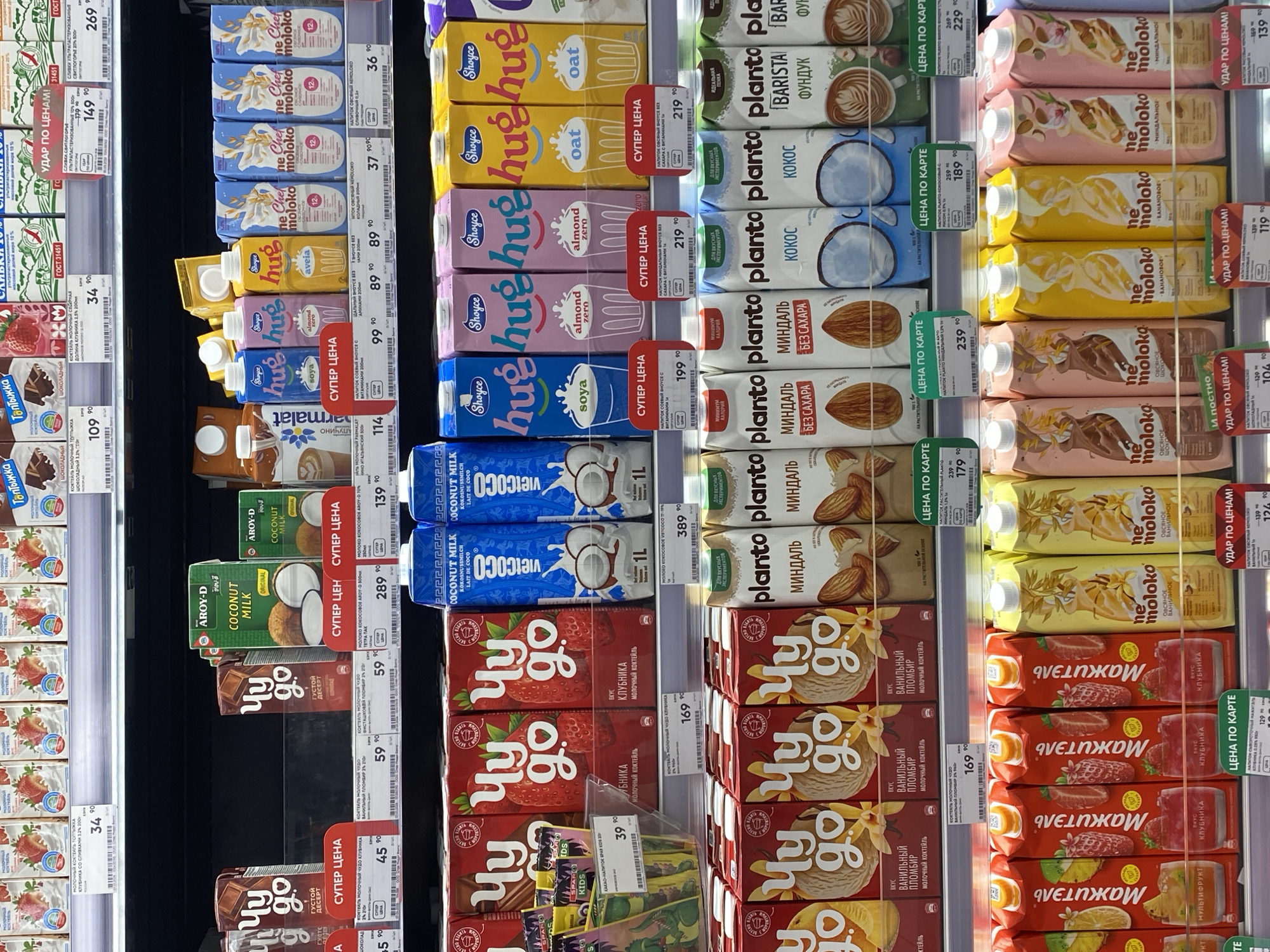 The image depicts a refrigerated shelf in a grocery store in Kazan, Russia. The shelves are stocked with a variety of milk and milk alternatives. Several brands are visible, including "Chudo," "Aroy-D," "Parmalat," "Vietcoco," "Shoyce," "Planto," and "Ne Moloko." The products include flavored milk drinks, coconut milk, soy milk, almond milk, oat milk, and barista-style milk. Price tags in Russian rubles are displayed for each product. The lighting is bright, typical of a supermarket environment.