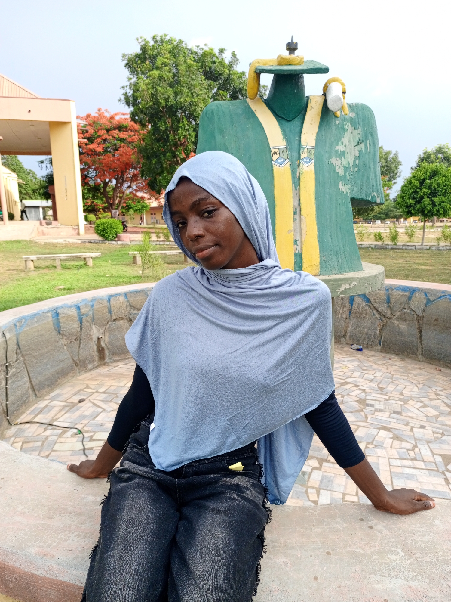 A young woman wearing a light blue hijab, a black long-sleeved top, and dark-wash jeans sits on a low, stone-tiled wall. Behind her is a sculpture of a person wearing a green robe with yellow details. The sculpture stands on a stone base. In the background, there are trees, grass, a building with a tan facade, and a red-flowered tree. The location appears to be a park or campus. The city and region are not identifiable from the image.