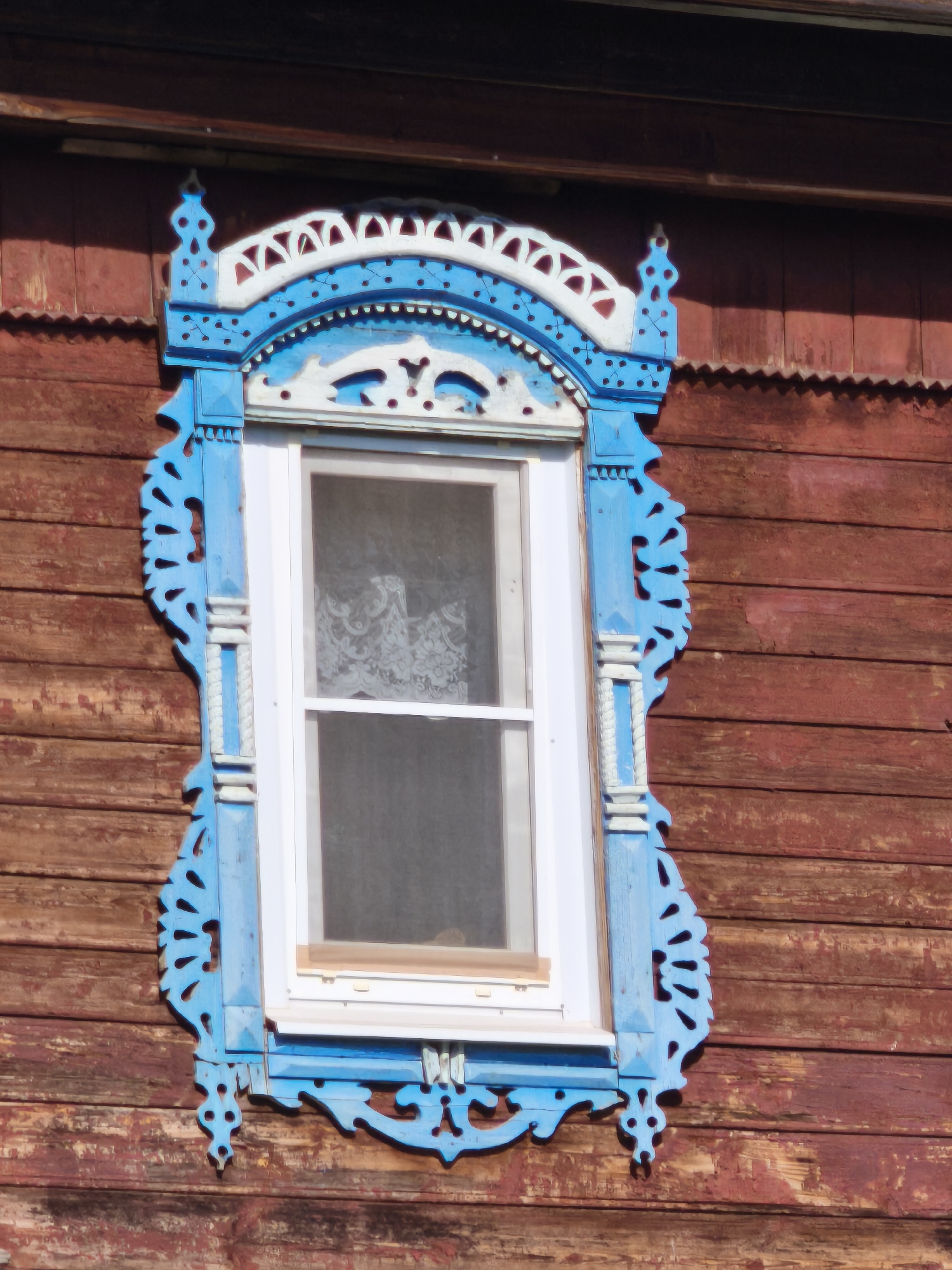 The image shows a close-up, eye-level view of a wooden window frame decorated with intricate blue and white carved wooden trim. The window itself is white-framed and appears to be made of glass with a white lace curtain visible inside. The window is set into a weathered wooden wall, painted a dark reddish-brown. The trim is ornate, featuring geometric patterns, stylized floral motifs, and decorative elements that extend from the sides and top of the window. The wood of the wall is aged, with visible grain and some signs of wear. The lighting suggests it is daytime, with sunlight casting shadows and highlighting the texture of the wood. The image focuses solely on the window and its immediate surroundings, giving no indication of the broader environment or location beyond the architectural style, which is common in rural Russian architecture.