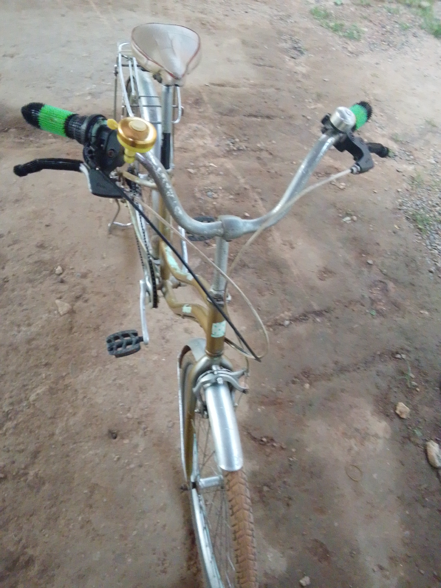 The image shows a close-up, overhead view of a bicycle parked on a dirt path in Serti, Nigeria. The bicycle has a light-colored seat and a metallic gold frame. The handlebars are silver, with green grips on either side. A brass bell is mounted on the right handlebar. The front wheel, fender, and a portion of the front tire are visible. The bicycle is positioned on a ground surface composed of dirt and small stones, with sparse, dry vegetation visible in the background. The lighting suggests it is daytime.