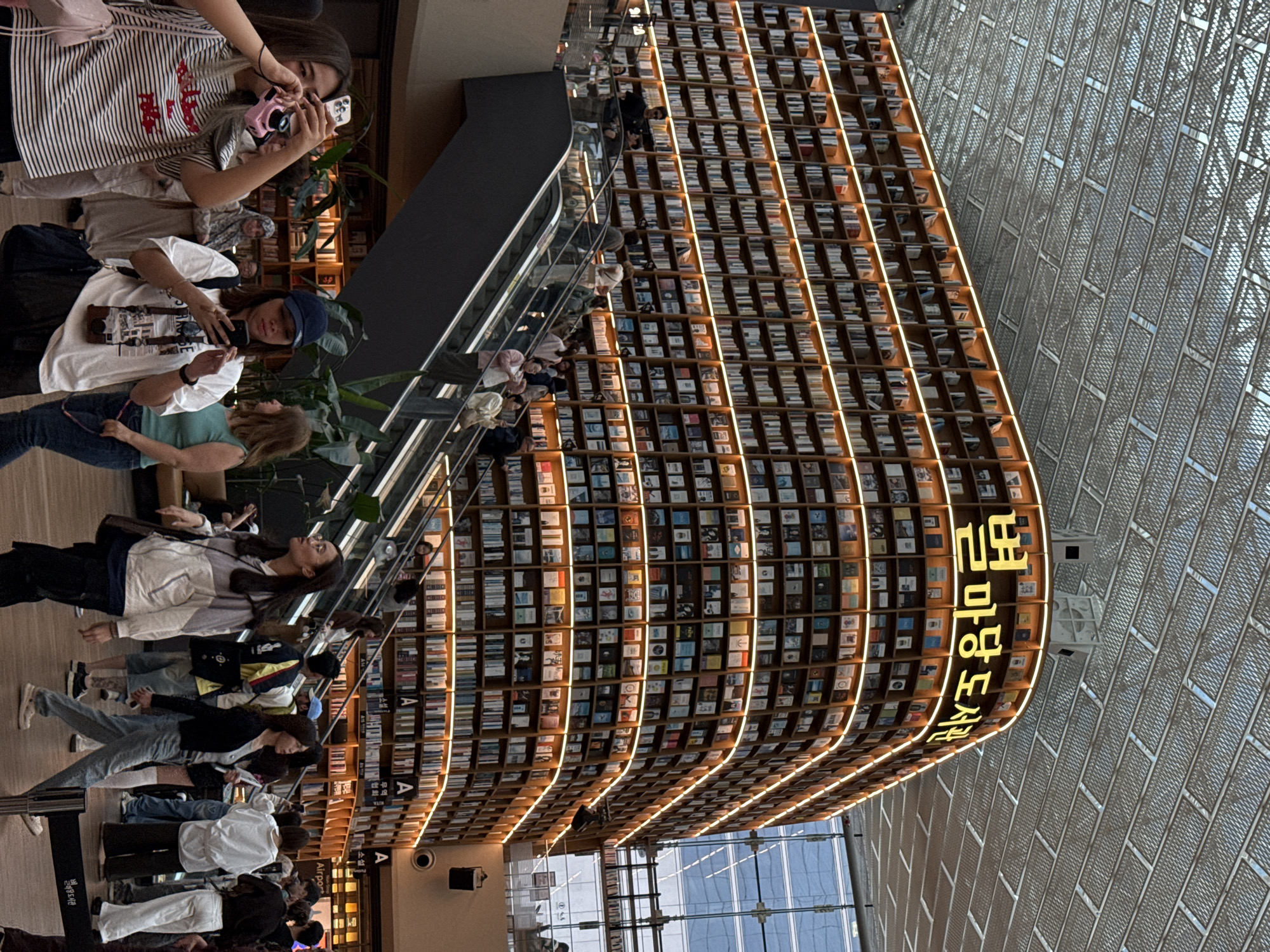 The image shows the Starfield Library in Seoul, South Korea, a large public space with floor-to-ceiling bookshelves filled with books. People are seen walking around and taking pictures. The architecture includes a modern, open design with a grid-like ceiling. A staircase with a glass barrier runs alongside the bookshelves. The library is brightly lit, and there is a large illuminated sign in Korean visible on one end of the bookshelves.