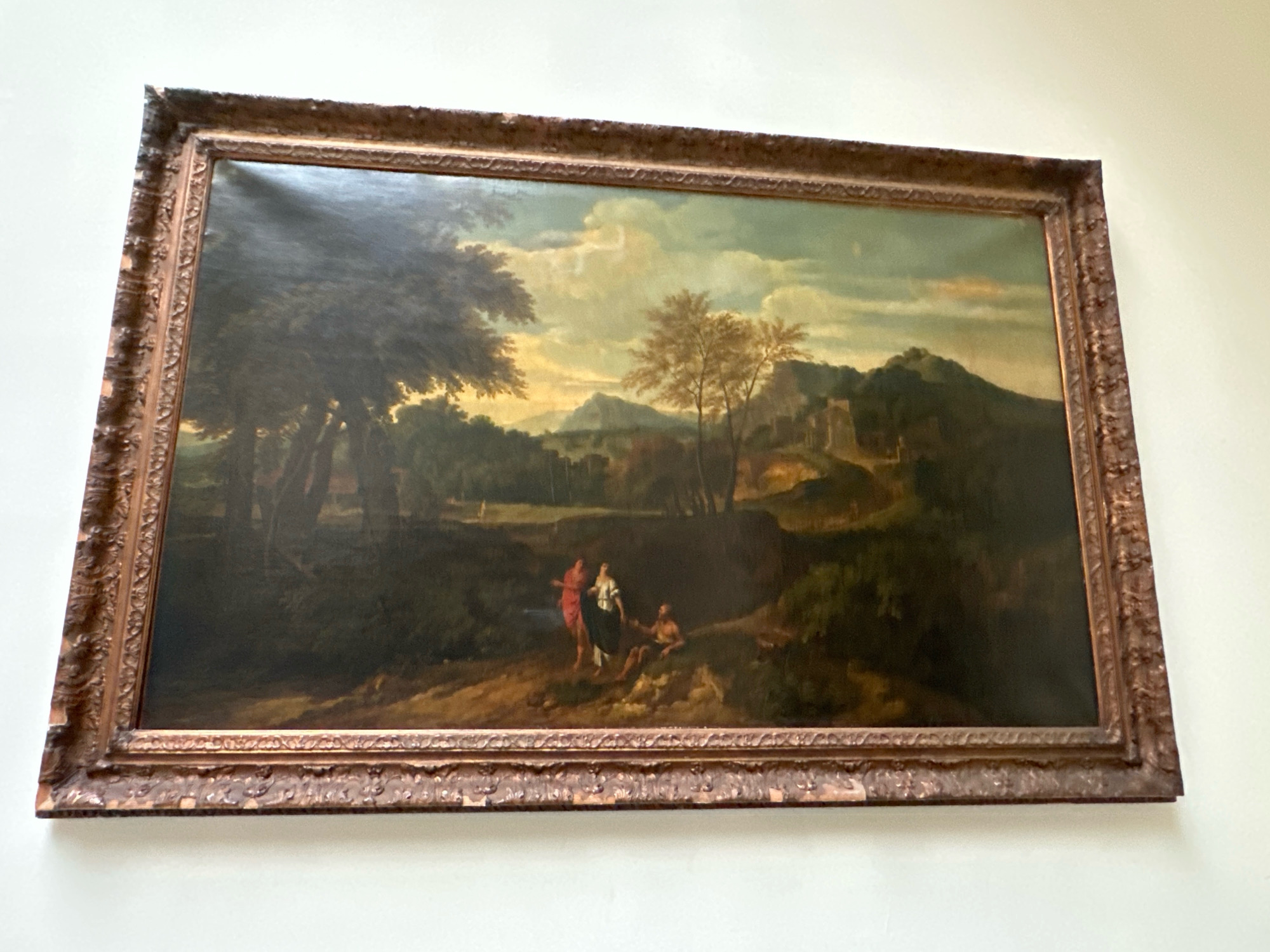 This is a horizontal, framed oil painting depicting a landscape with three figures. The painting is hung on a plain, light-colored wall. The frame is ornate and appears to be made of gilded wood with intricate carvings.
The landscape is lush and verdant, with dark trees dominating the left side of the painting and a lighter, more open area stretching towards the right. In the distance, rolling hills and mountains can be seen, with a suggestion of a building or ruins on one of the hills. The sky is a blend of blue and pale yellow, with clouds that suggest either dawn or dusk.
In the foreground, on a dirt path, are three figures. Two are standing, and one is seated. The standing figures appear to be a woman in a red garment and another in a white and dark garment. The seated figure is a male, partially obscured. Their interactions are not clearly discernible from the image.
The painting is located in the National Museum of Serbia, in Belgrade.