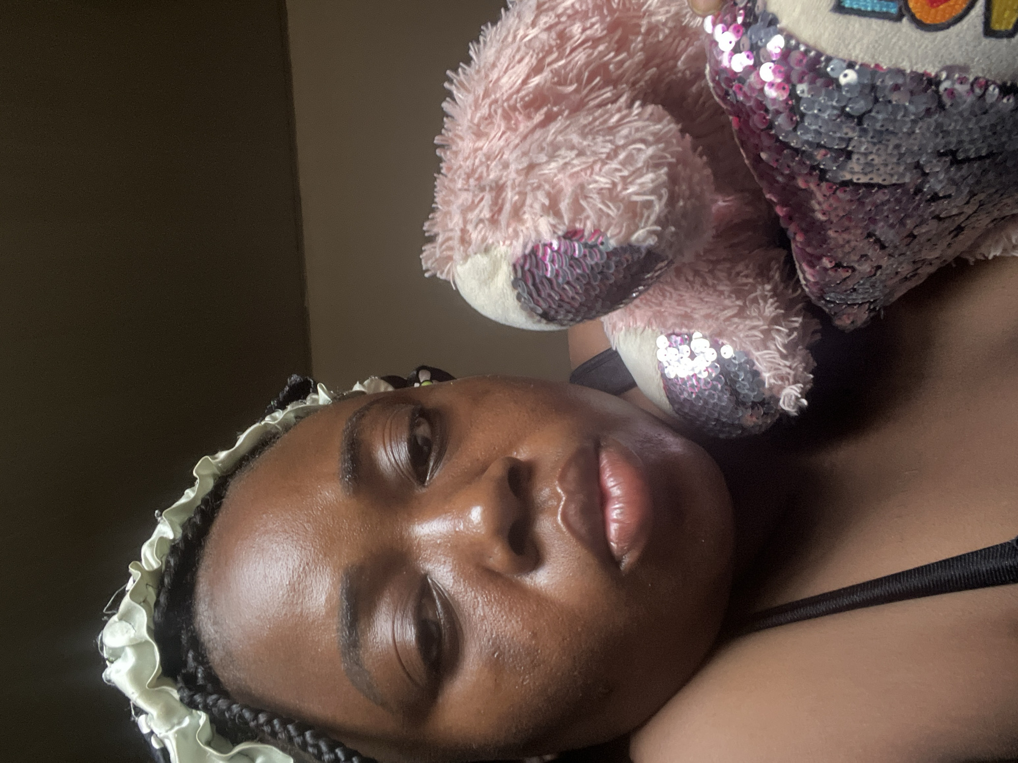 The media file presents a rotated close-up portrait of a young Black woman, likely in an indoor setting in Abuja, Nigeria. She is lying down, looking directly at the viewer. Her skin appears radiant with highlights from direct lighting, and her lips are slightly parted and glossy. She wears a light-colored, ruffled bonnet or sleep cap over her braided hair.
She is holding or cuddling a plush pink teddy bear, parts of which are adorned with reversible silver and pink/purple sequins. A prominent feature on the teddy bear is a heart-shaped sequined patch that displays the colorful word "LOVE". The background is a plain, dark wall, suggesting a simple room. The strong lighting creates bright highlights on her face and the teddy bear, emphasizing a relaxed and personal moment. A black bra strap is visible on her shoulder. The overall impression is one of a calm, intimate daytime scene.