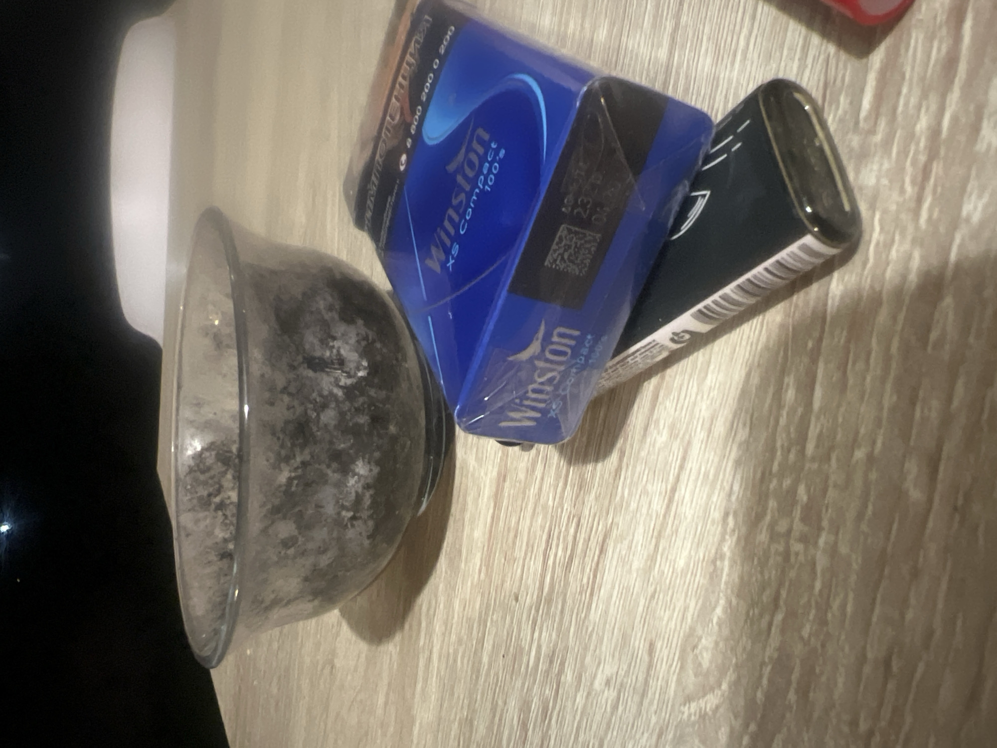 A medium shot captures a wooden table with a glass bowl filled with a dark, granular substance. To the right of the bowl, a blue pack of Winston XS Compact 100's cigarettes rests on top of a black lighter, which has a metallic gold trim and a white striped pattern along its side. The scene is dimly lit, suggesting an indoor setting at night. The background is dark, with a faint blurry light visible in the upper left corner. The table surface exhibits a light wood grain pattern. There is no indication of the city of Krasnye Orly, Russia, or any people in this image.