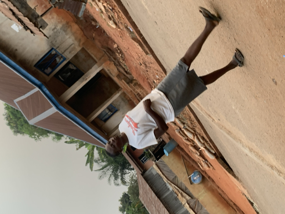 A man wearing a white t-shirt and gray shorts stands on a dirt road. He has sandals on his feet. Behind him is a building, partially constructed, with exposed brick and visible windows. Green foliage surrounds the building, suggesting a natural setting. The area appears rural or developing, with a mix of modern construction and more traditional building styles.