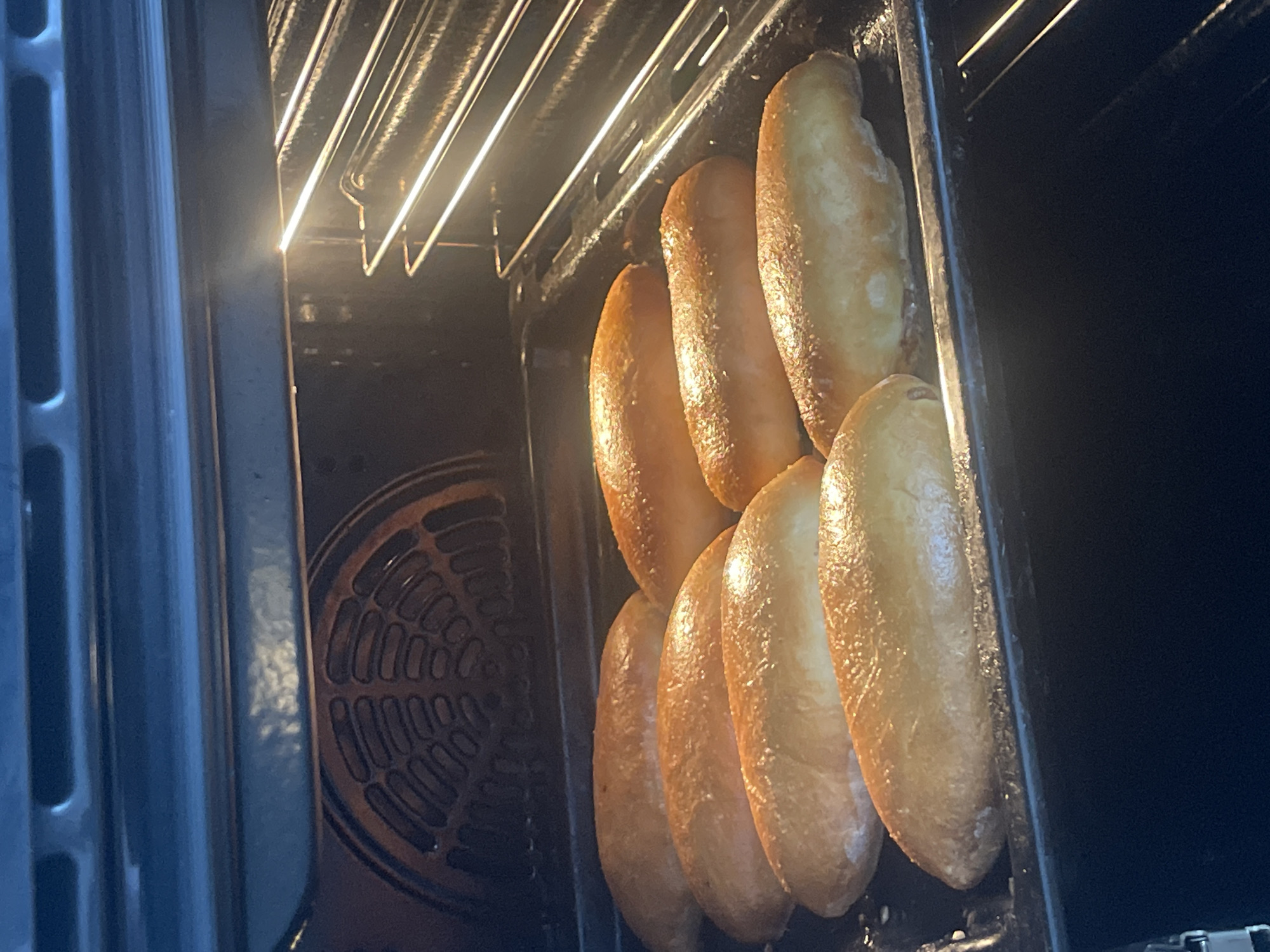 The image shows a row of six oblong-shaped, golden-brown baked goods on a black baking tray inside an oven. The oven light is on, illuminating the interior and the upper heating element. The oven door is slightly ajar. Details beyond the interior of the oven are obscured. The location (Izhevsk, Russia) is not verifiable from the visual information provided.