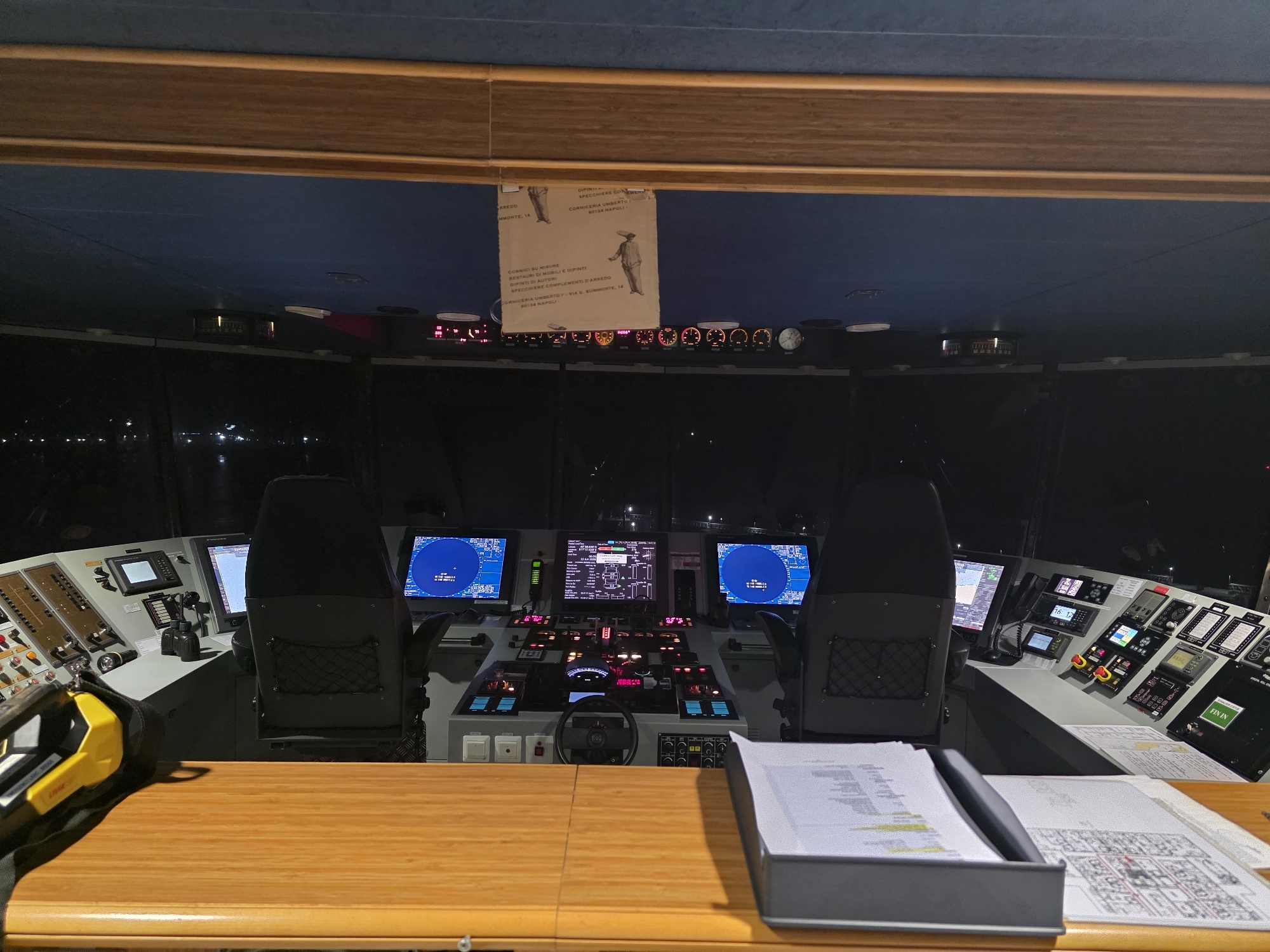 The image depicts the interior of a ship's bridge, likely at night. The view is from behind a wooden console, looking towards the front of the bridge. Several large screens display navigational information, including radar and electronic charts. There are two captain's chairs positioned in front of the consoles. Various control panels with buttons, switches, and gauges are visible. A yellow and black handheld device rests on the wooden console in the foreground. The background shows a dark expanse, suggesting open water, with distant lights possibly indicating a port or city. A document, possibly a manifest or chart, is laid out on the console. The overall impression is one of a functional and technologically advanced maritime control center. The presence of specific ship equipment suggests a vessel operating within or departing from a port. Given the context of Brindisi, Italy, this could be a ship at sea near the port of Brindisi, or docked within the port itself, preparing for departure or having just arrived.