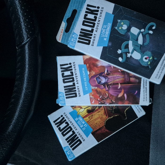 Three boxes of Unlock! Short Adventures games are displayed. The topmost box, "Le Chat de M. Schrödinger," features cartoon cats. Below it, "Le Vol de L'Ange" shows a hooded figure, and the bottom one, "Le Réveil de la Momie," depicts a mummy-themed image. Each box is a white rectangle with a blue banner containing the game title. They are positioned diagonally against a dark background. The wheel of a vehicle is visible on the side.