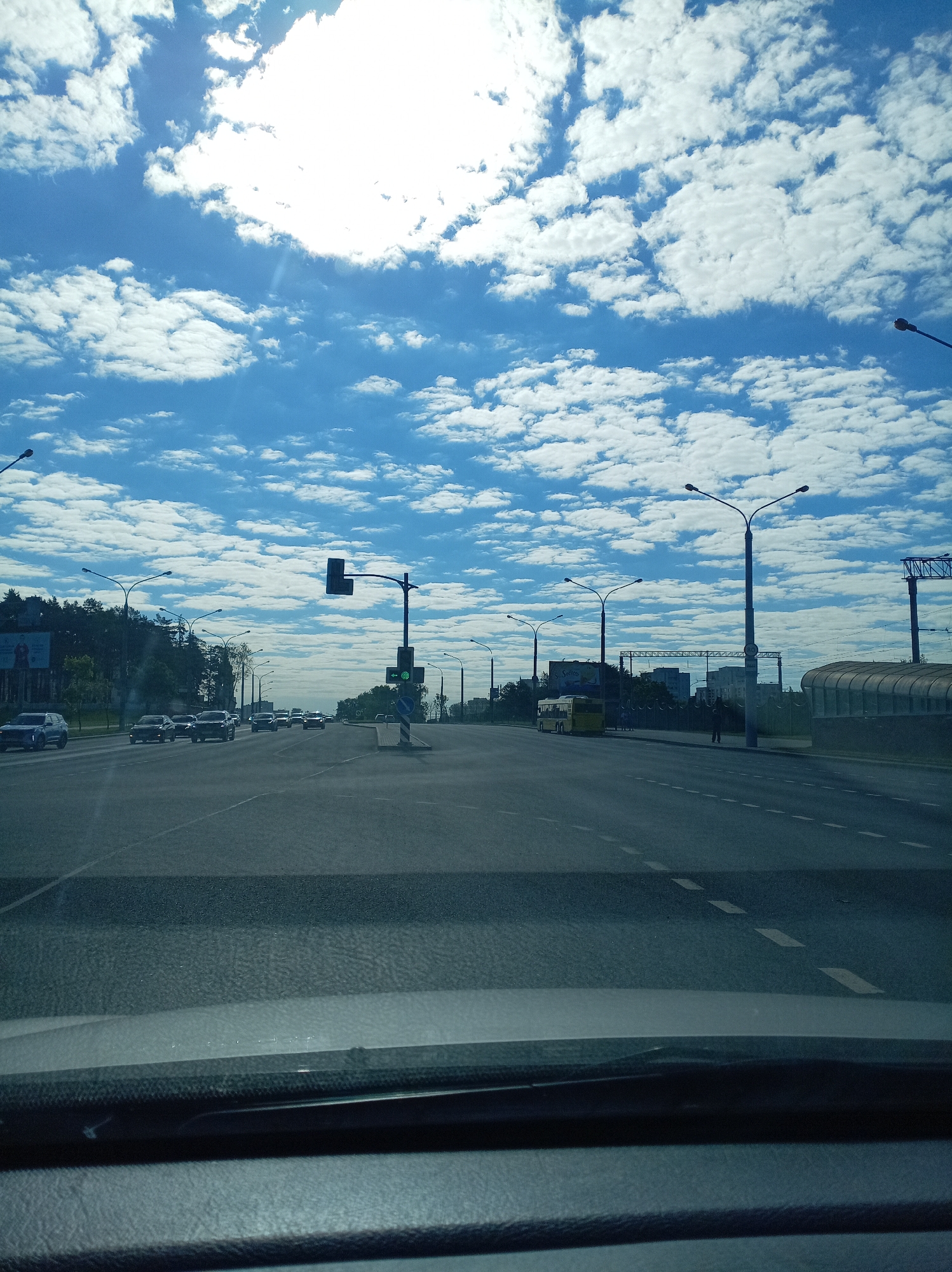 The image presents a daytime view of a multi-lane asphalt road from the perspective inside a vehicle, with the dashboard visible at the bottom. The upper half of the frame is dominated by a bright blue sky filled with numerous scattered white cumulus clouds. The sun is partially obscured by a large cloud in the upper left, creating a prominent glare.
Several cars, mostly dark-colored sedans or SUVs, are visible on the road, with some moving and others appearing stationary. White dashed lines mark the traffic lanes. Numerous street lights with double arms line the road on both sides. A traffic light pole on the left displays a green light, indicating that traffic flow is permitted. A blue circular street sign is also visible near the traffic light.
On the left side of the road, a dense line of green trees forms a background, partially obscuring distant buildings. A large billboard is visible behind the trees, with some indistinct text possibly beginning with "C**bo." On the right side of the road, further down, a yellow bus is parked, bearing text that appears to be "Sohou" on its side. One person is vaguely discernible standing near the bus. A covered pedestrian structure or bus stop enclosure is also present on the far right.
The scene depicts an urban or suburban environment with active road traffic under clear, sunny weather conditions.