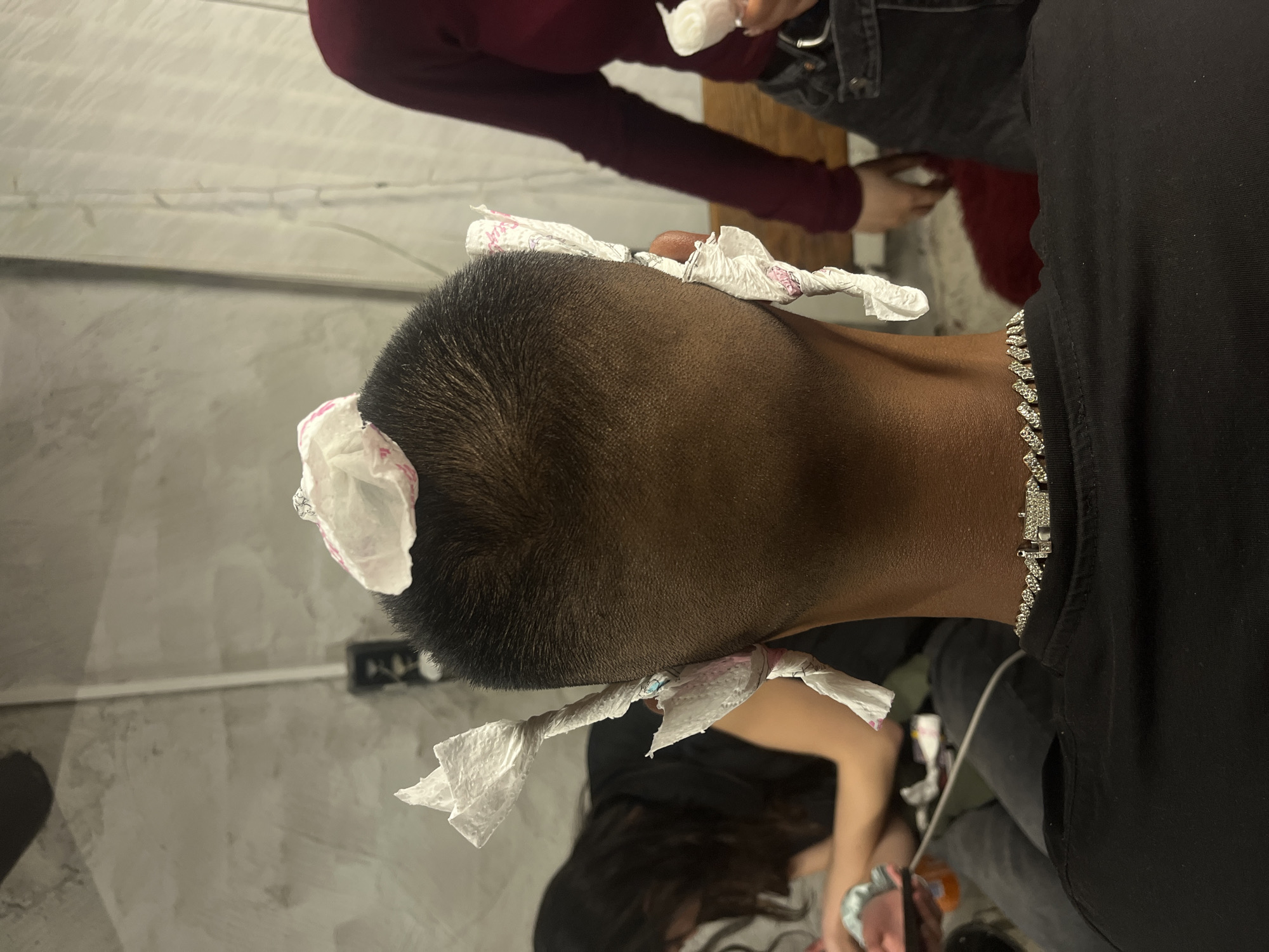 The image depicts a person with a short, dark haircut, and several pieces of white tissue paper attached to their head. They are wearing a black shirt and a silver chain necklace. The surrounding people are partially visible, suggesting that the focus is on the person with the tissue paper. There are no identifiable landmarks indicating a location in Lozarevo, Bulgaria.