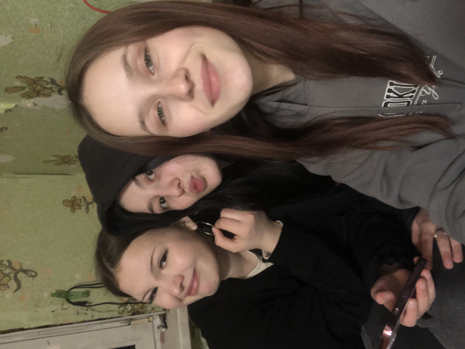 A selfie captures three young females in an indoor setting located in Foki, Russia. The individual on the left, with light brown hair, smiles subtly while holding a silver hoop earring with her right hand and a dark-colored smartphone with her left. She wears a dark long-sleeved top over a light undershirt. The central figure, with dark hair and wearing a black hooded sweatshirt with the hood up, makes a pouting or 'kiss' face. The individual on the right, with auburn hair, smiles broadly and wears a grey hooded sweatshirt featuring a partial white graphic print. The background consists of a light green wall covered in worn wallpaper with a repeating yellow-brown floral pattern, showing areas of peeling. A white-framed window is partially visible on the left, and a green object hangs on the wall near it. A red cable is partially visible on the far right.