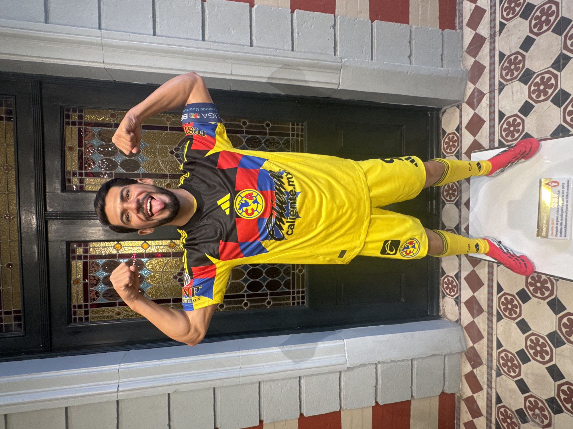 The image captures a man posing in front of a doorway in Mexico City, Mexico. He is wearing a yellow and black Club América soccer jersey with matching yellow shorts and socks. The jersey features the logos of Adidas and Caliente.mx, and the sleeves have the Liga BBVA logo. His red soccer cleats are visible, and he is flexing both arms with clenched fists while sticking out his tongue.
The doorway is painted black and has decorative stained glass windows. The walls around the doorway are made of grey brick with a red and white tile pattern at the bottom. The floor is covered in patterned tiles with a geometric design. To the right of the man's foot, a small plaque with text is visible.
