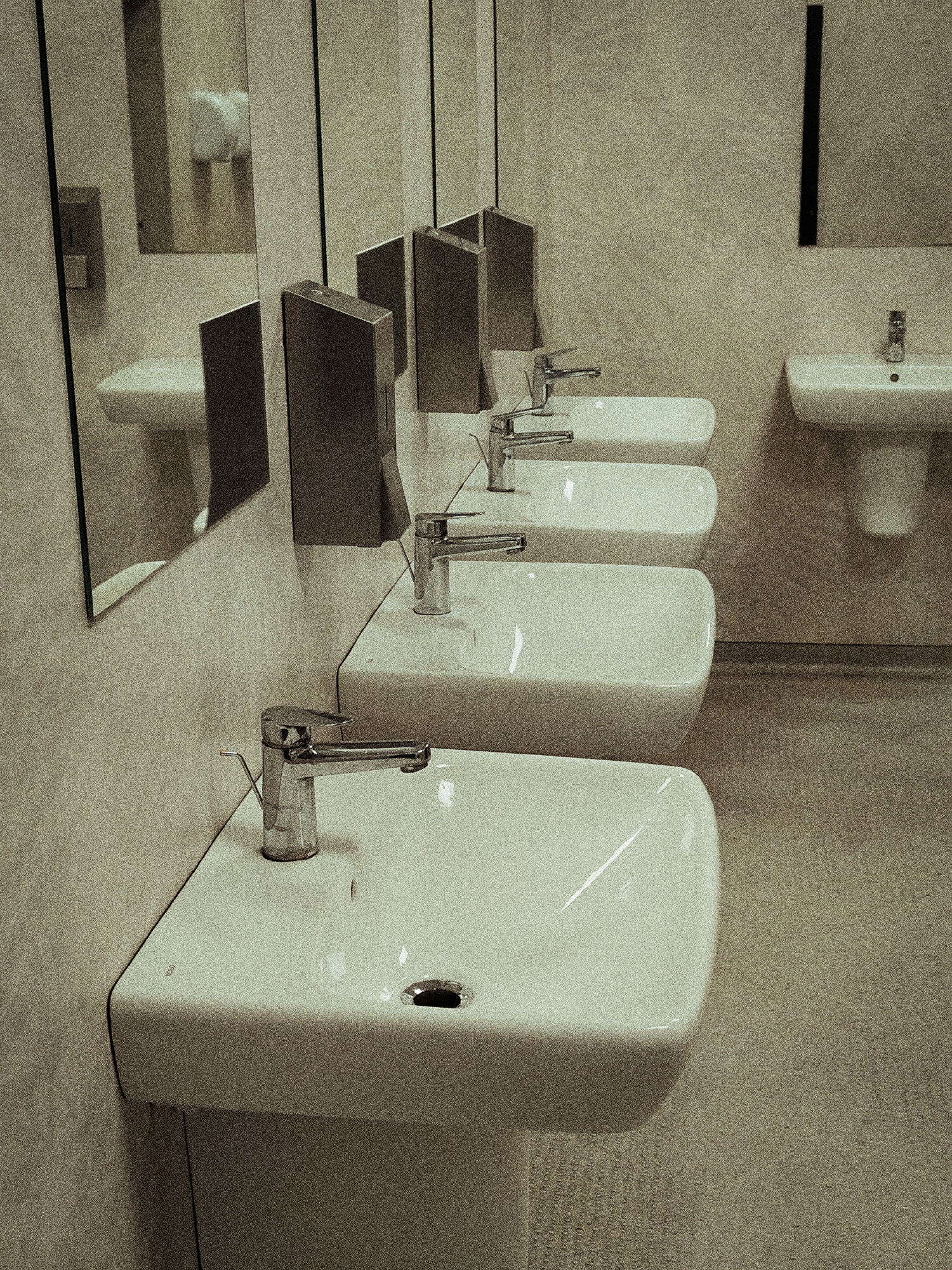 The image depicts a row of white ceramic sinks with chrome faucets in a public restroom. The sinks are modern and rectangular, and the faucets are of a lever-operated design. The environment appears clean and well-maintained, with tiled walls and a speckled floor. Mirrors are present, reflecting the sinks and the restroom's lighting. The scene is static, with no people visible. The overall impression is one of a functional and minimalist space. This restroom is located in the city of Łomna-Las, Poland.